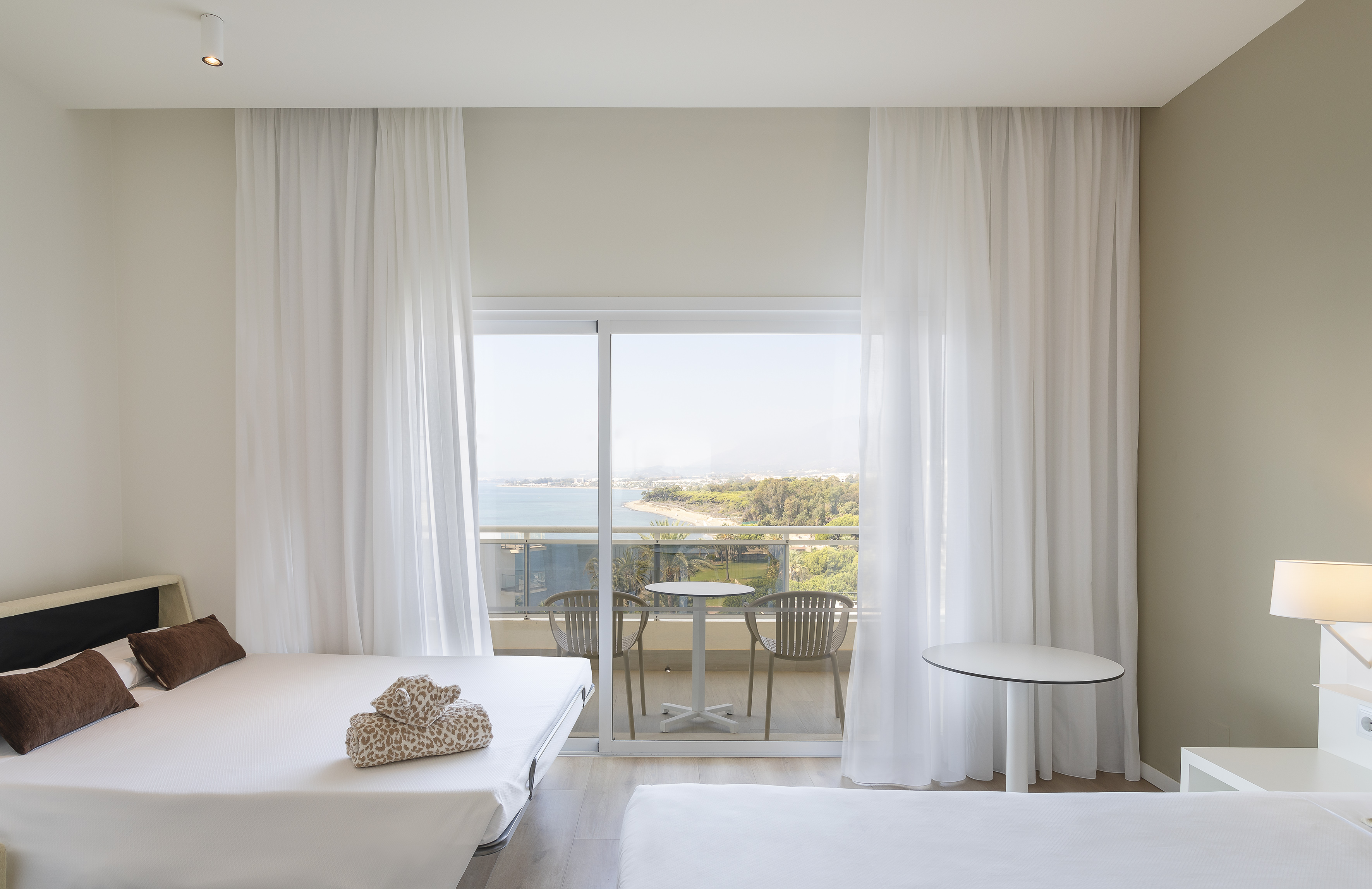 Bright hotel room with ocean view balcony, two beds, and white sheer curtains.
