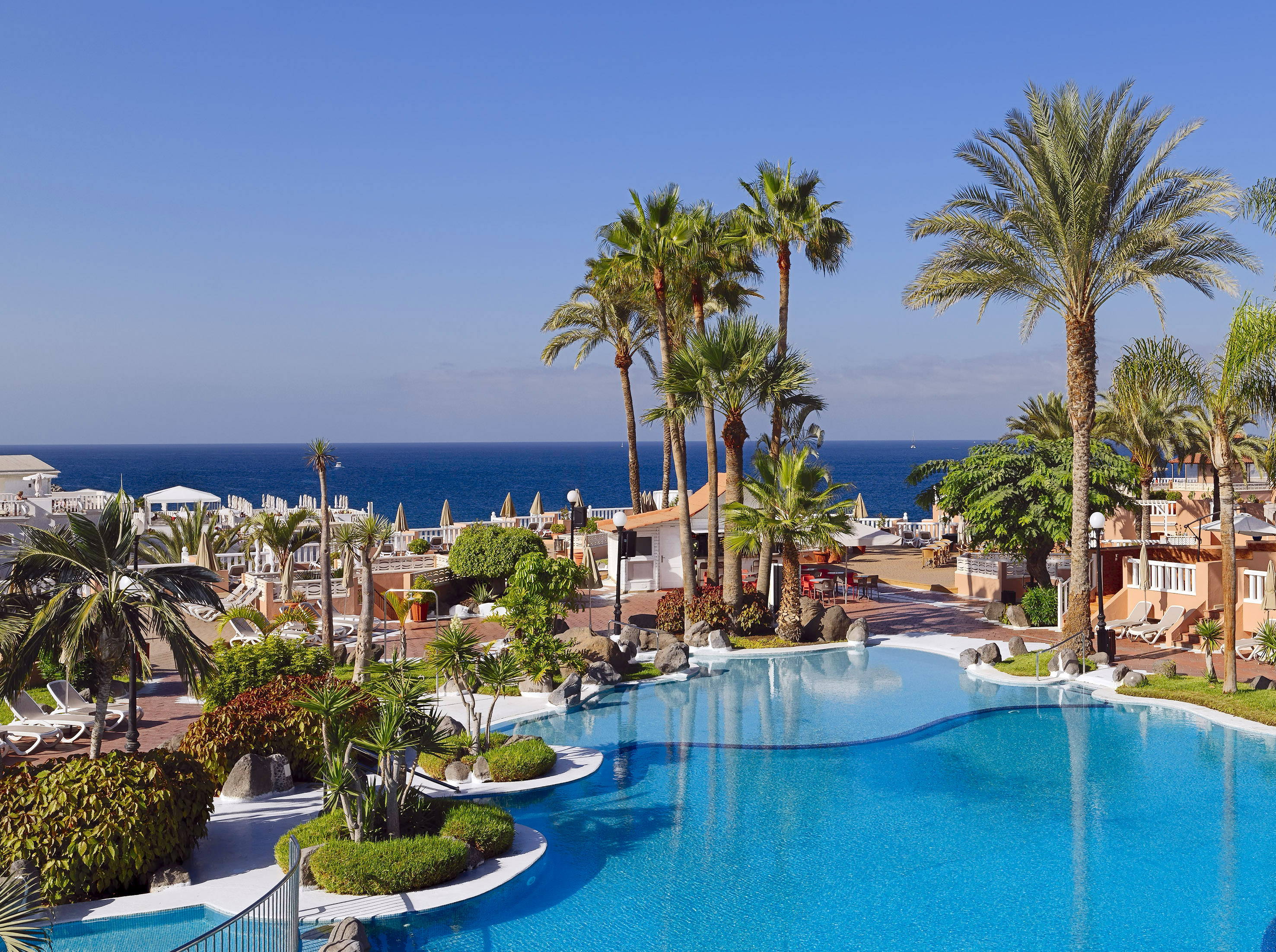 Tropical resort with large blue swimming pool, palm trees, and ocean view.