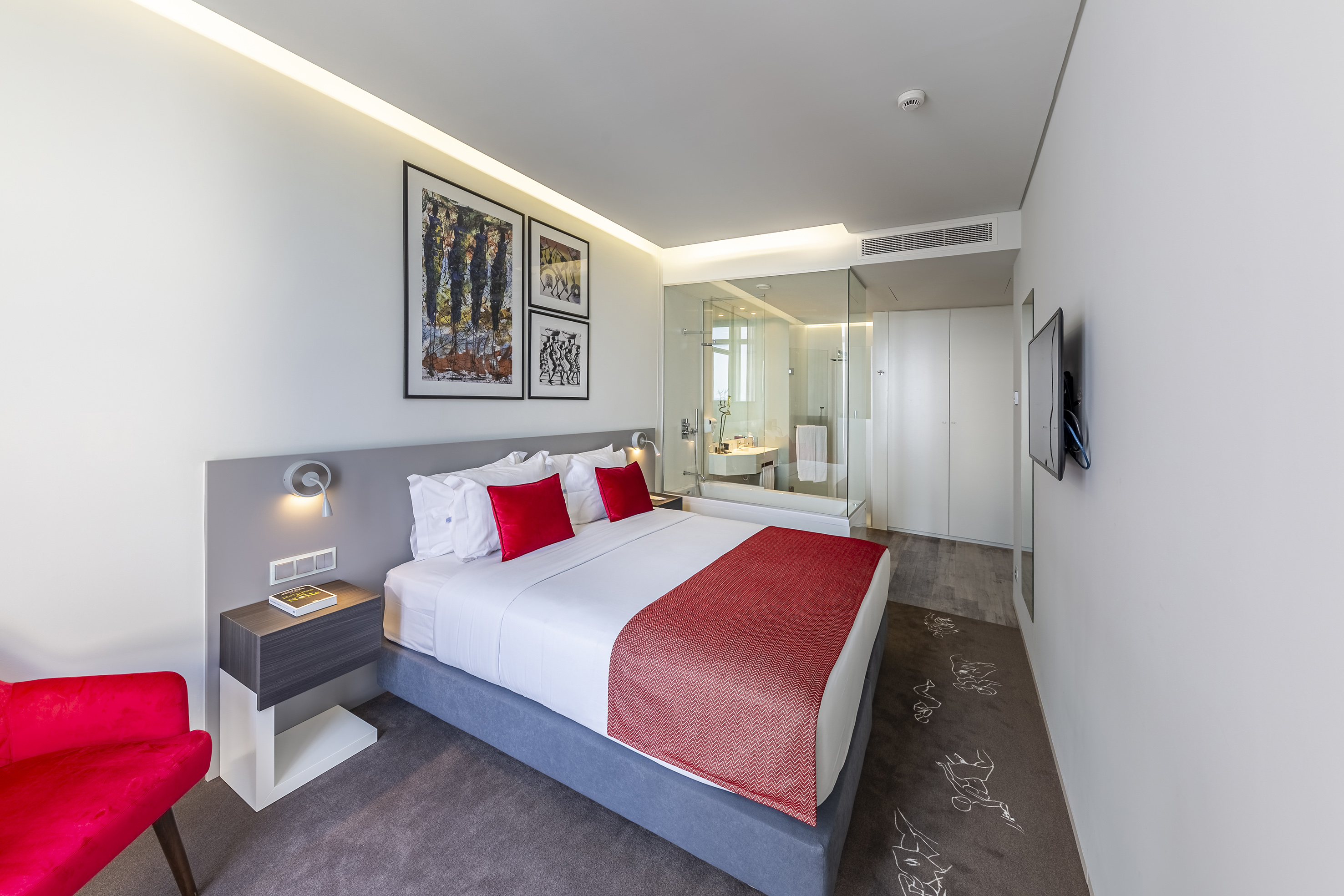 Modern hotel bedroom with king bed, red accents, glass shower, and wall TV.