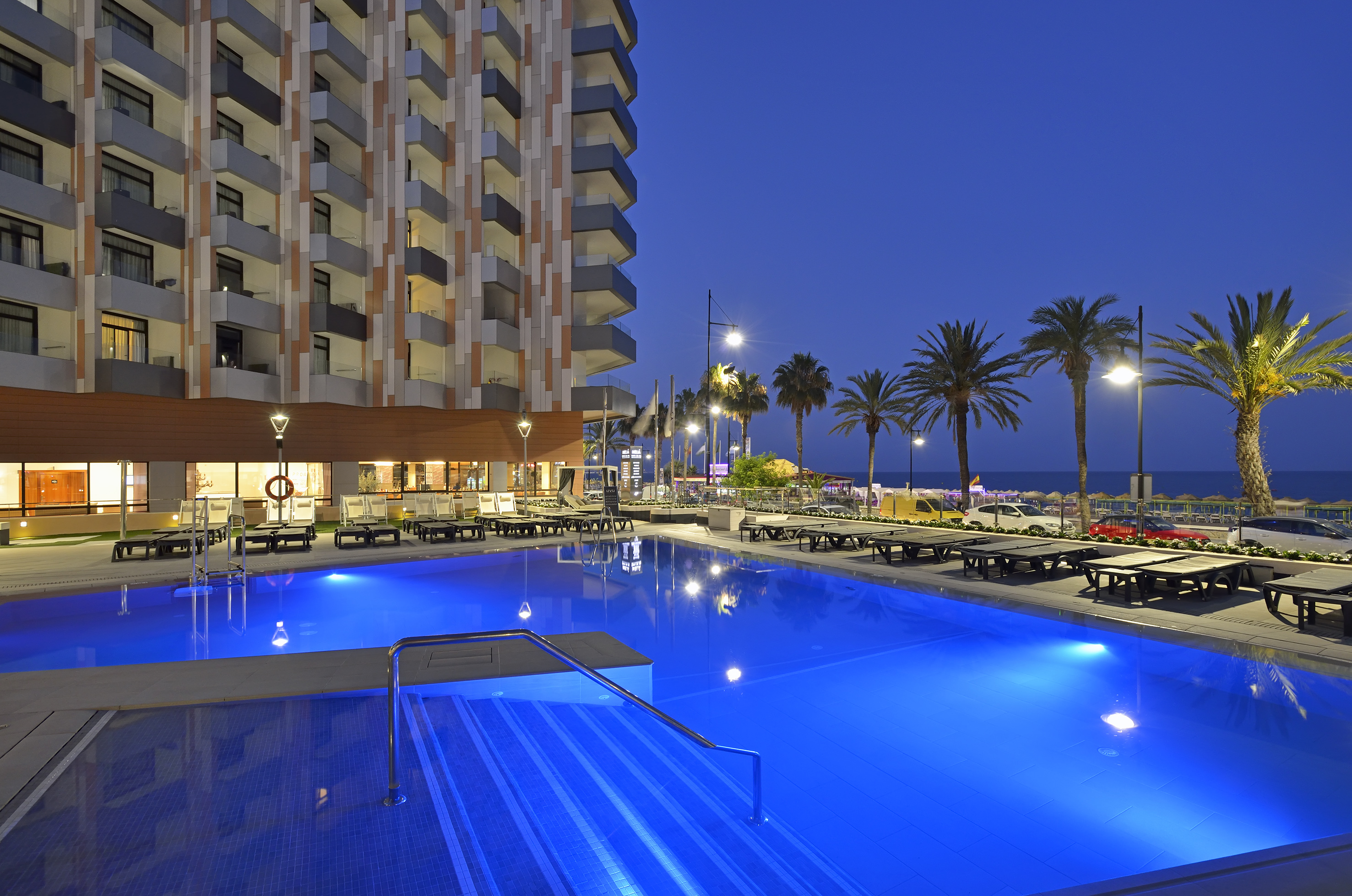 Modern hotel with illuminated blue swimming pool, palm trees, and ocean at dusk.