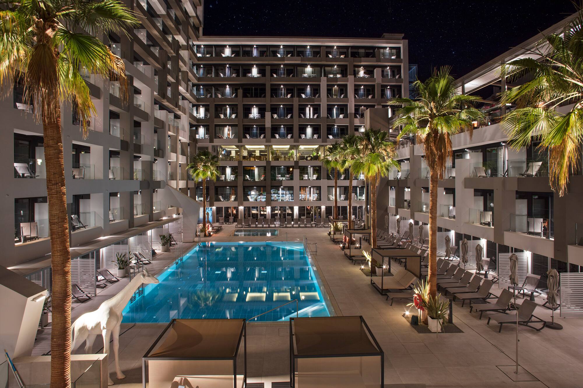 Illuminated modern hotel resort pool, palm trees, and giraffe statue at night.