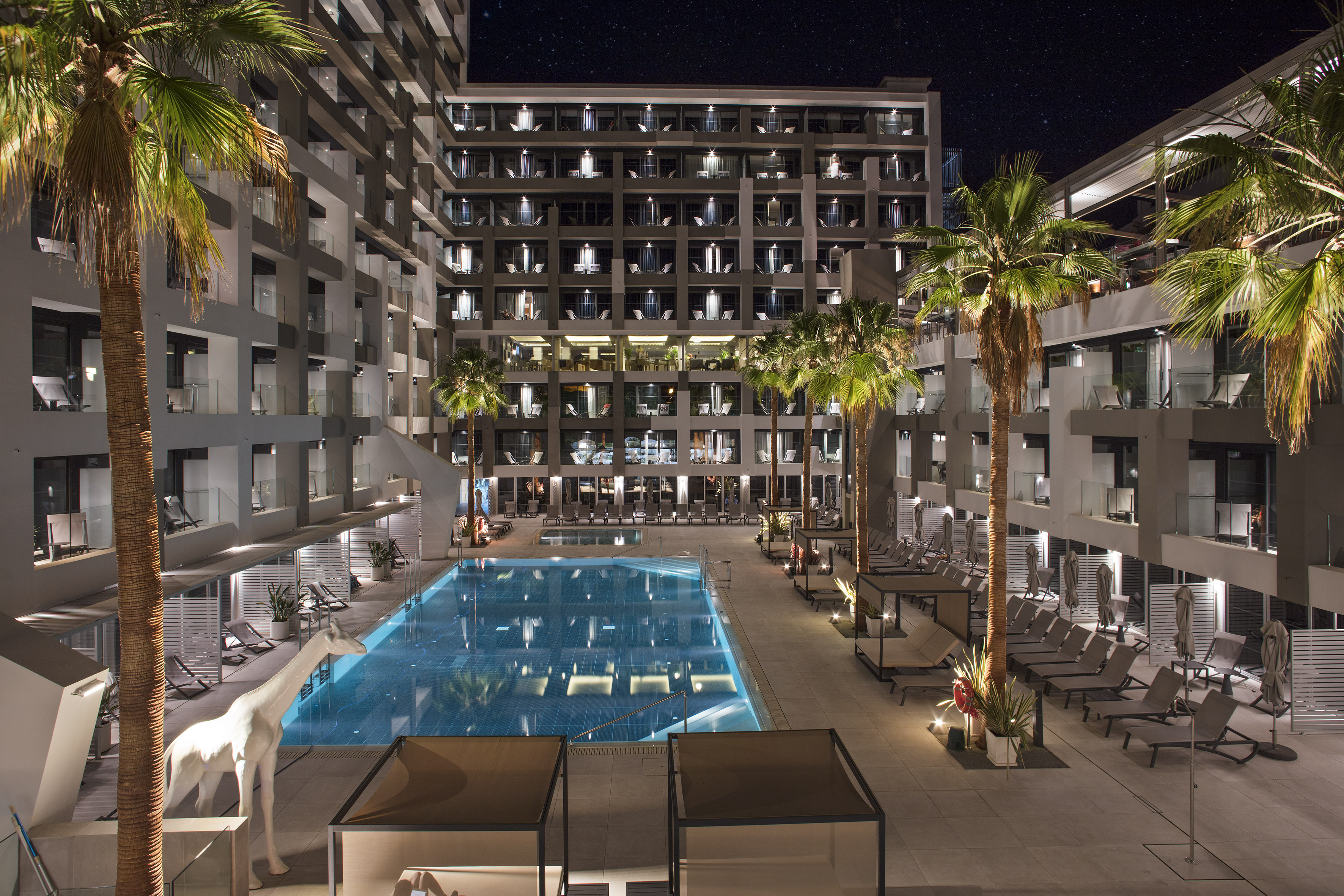 Illuminated modern hotel resort pool, palm trees, and giraffe statue at night.