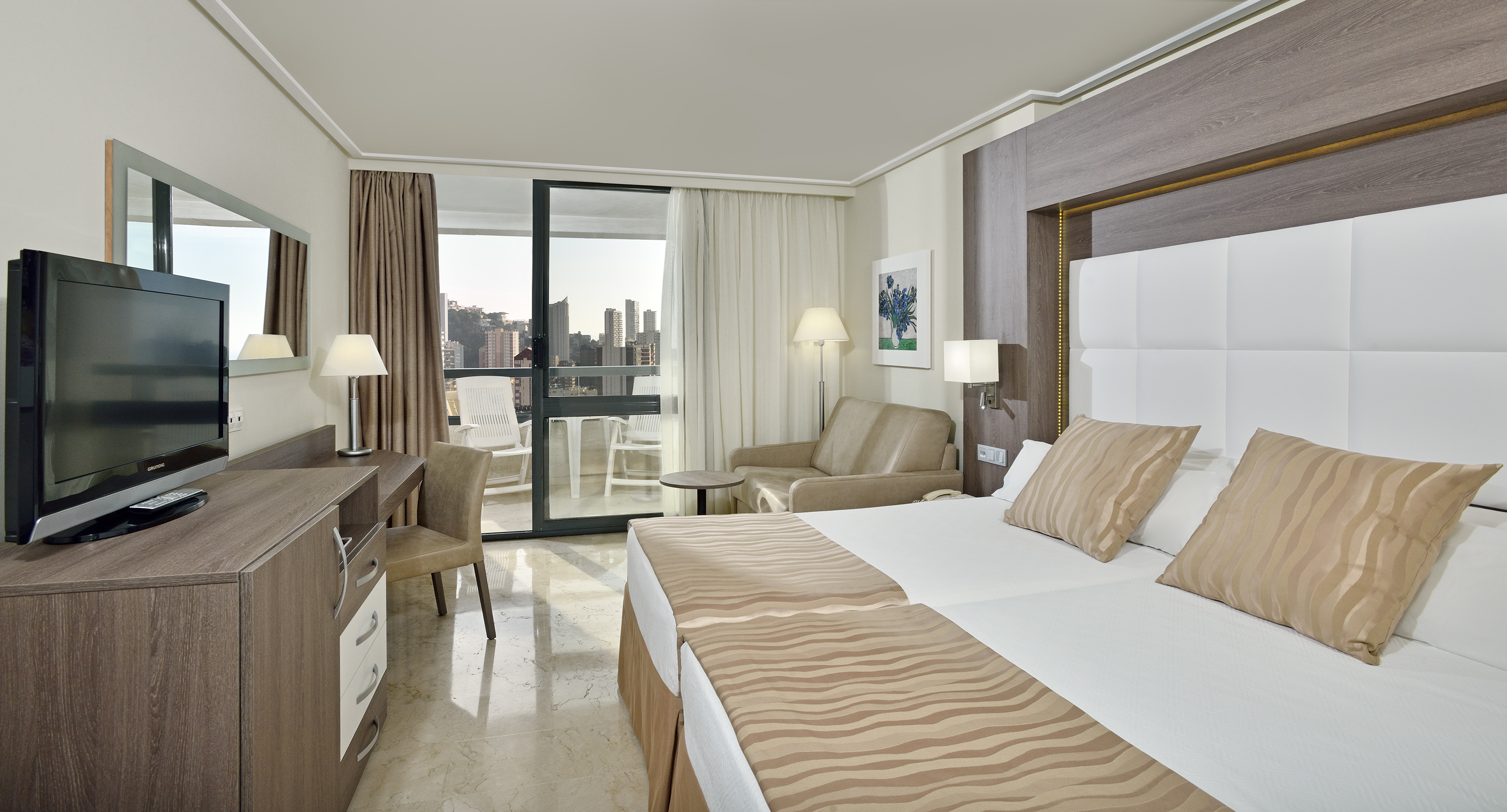Modern hotel room with twin beds, TV, balcony, and city skyline view.