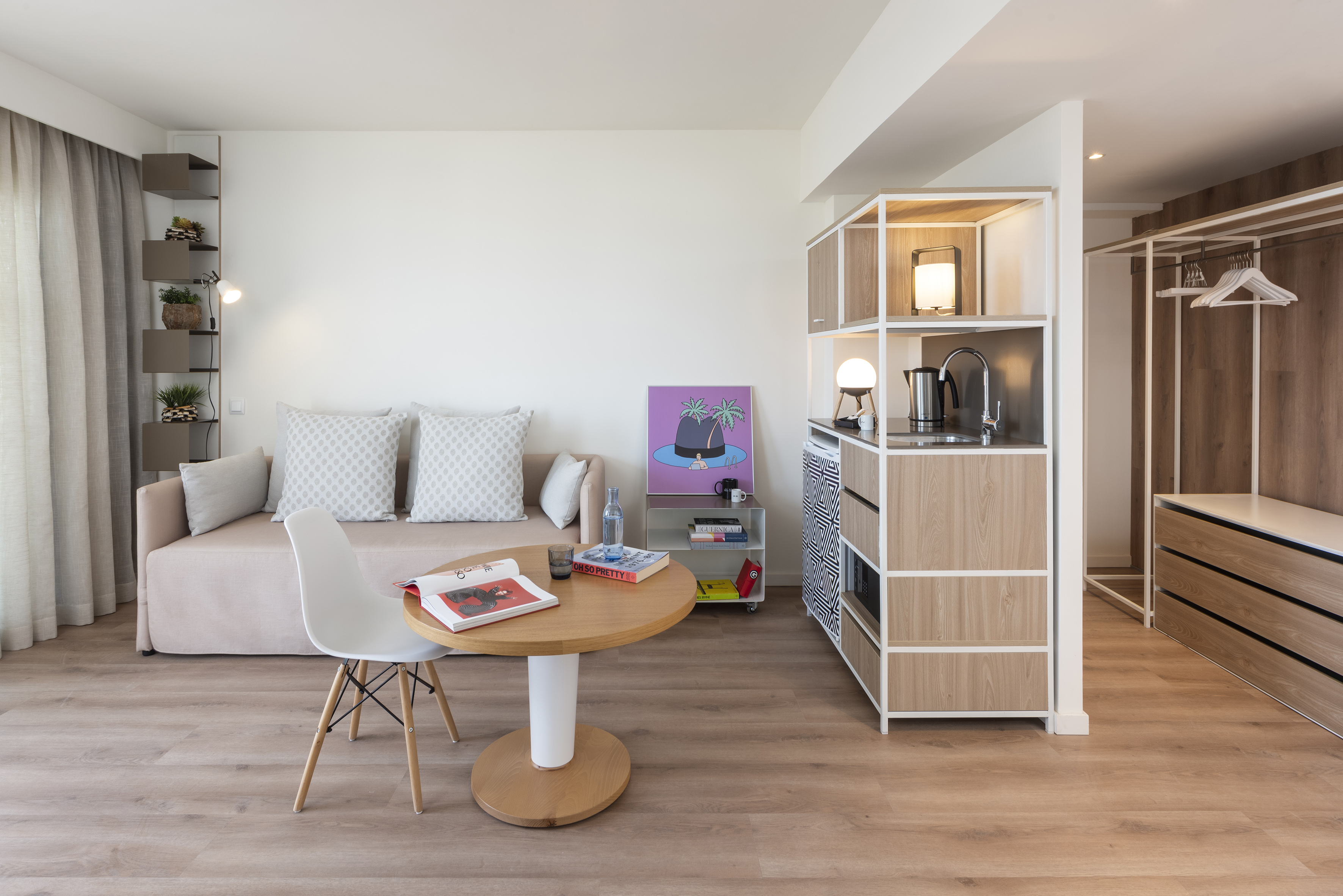 Modern studio apartment with sofa, dining table, compact kitchen, and open wardrobe.