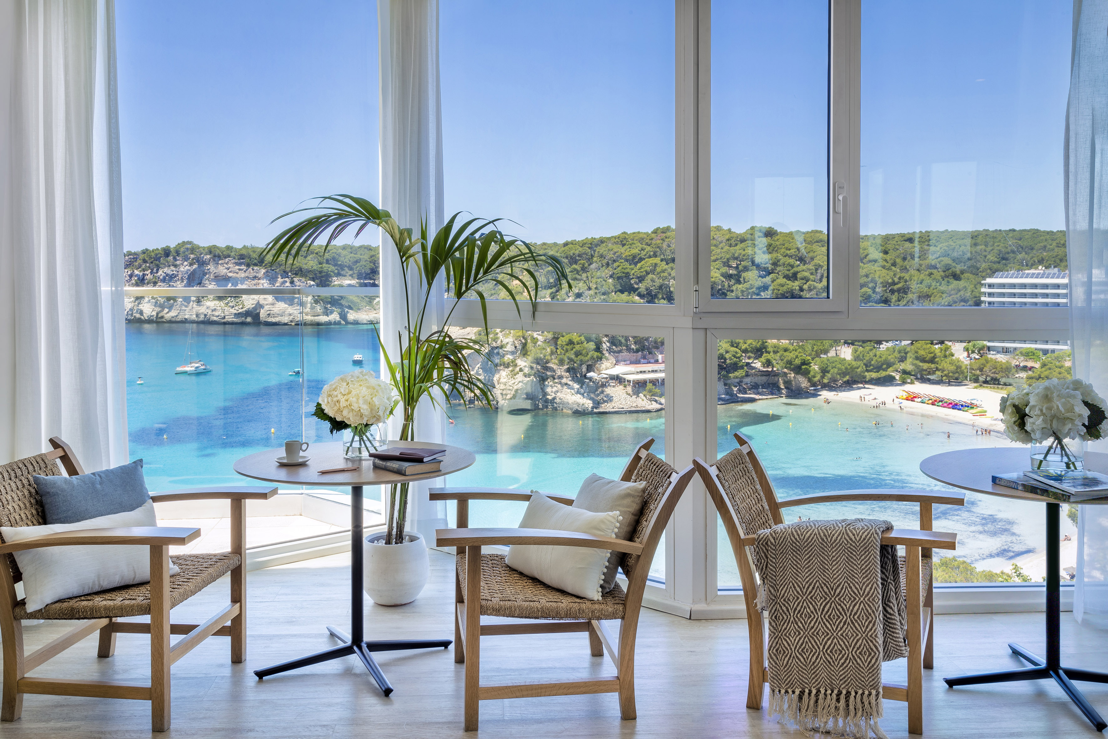Hotel room with chairs, table, overlooking sunny turquoise bay and sandy beach.