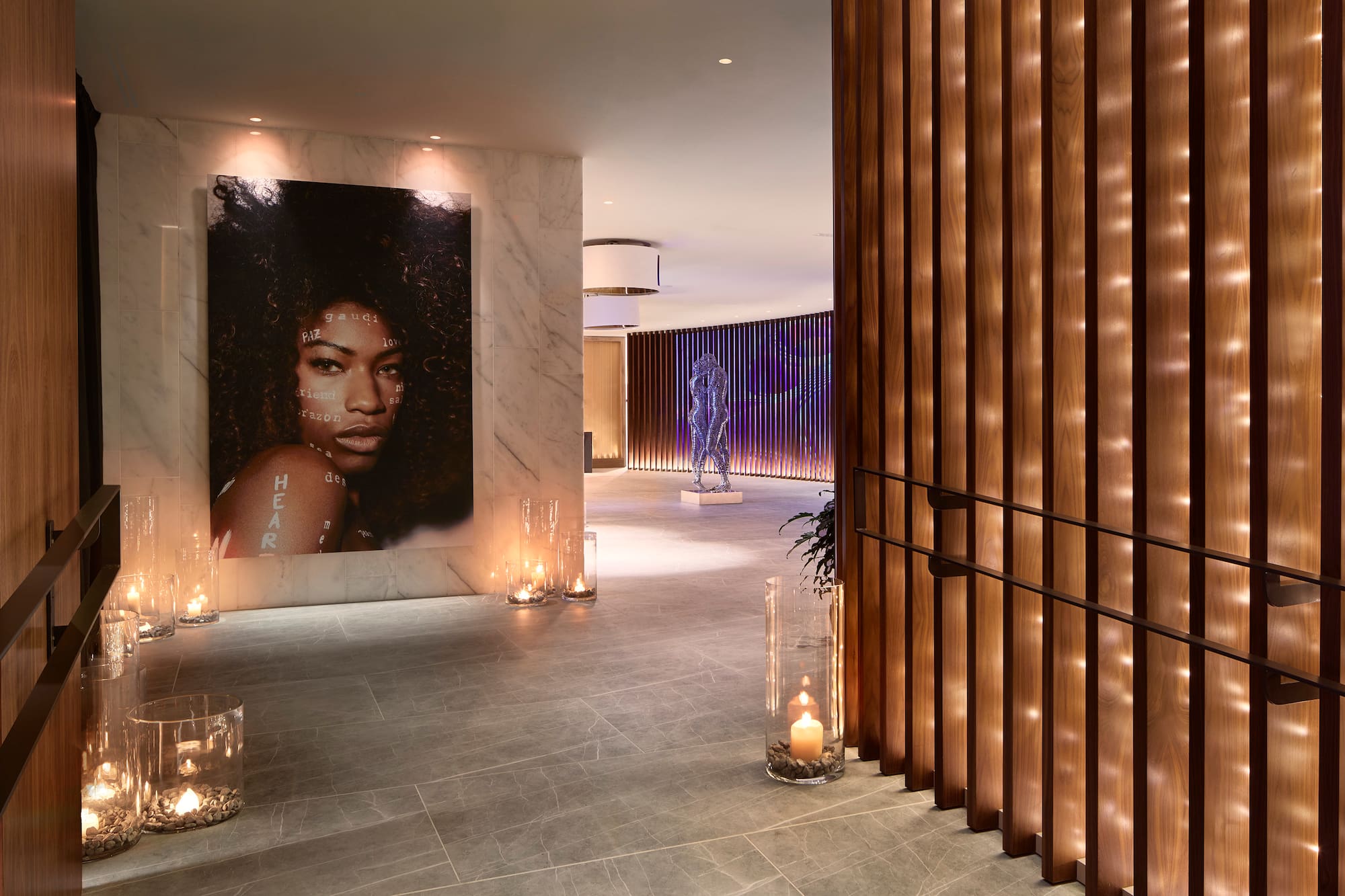 Modern hallway with portrait art, wood slats, sculpture, and ambient candles.