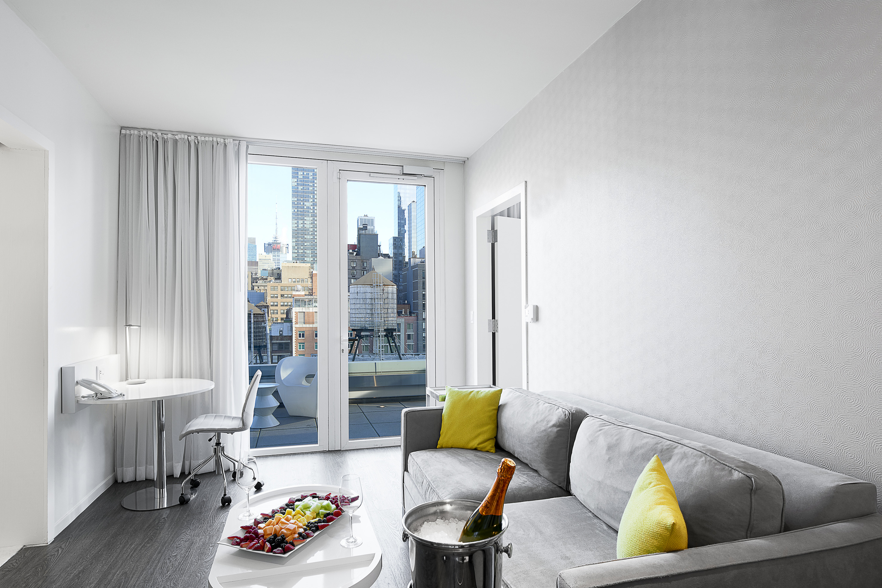 Modern hotel suite with city view balcony, gray sofa, champagne, fruit platter.