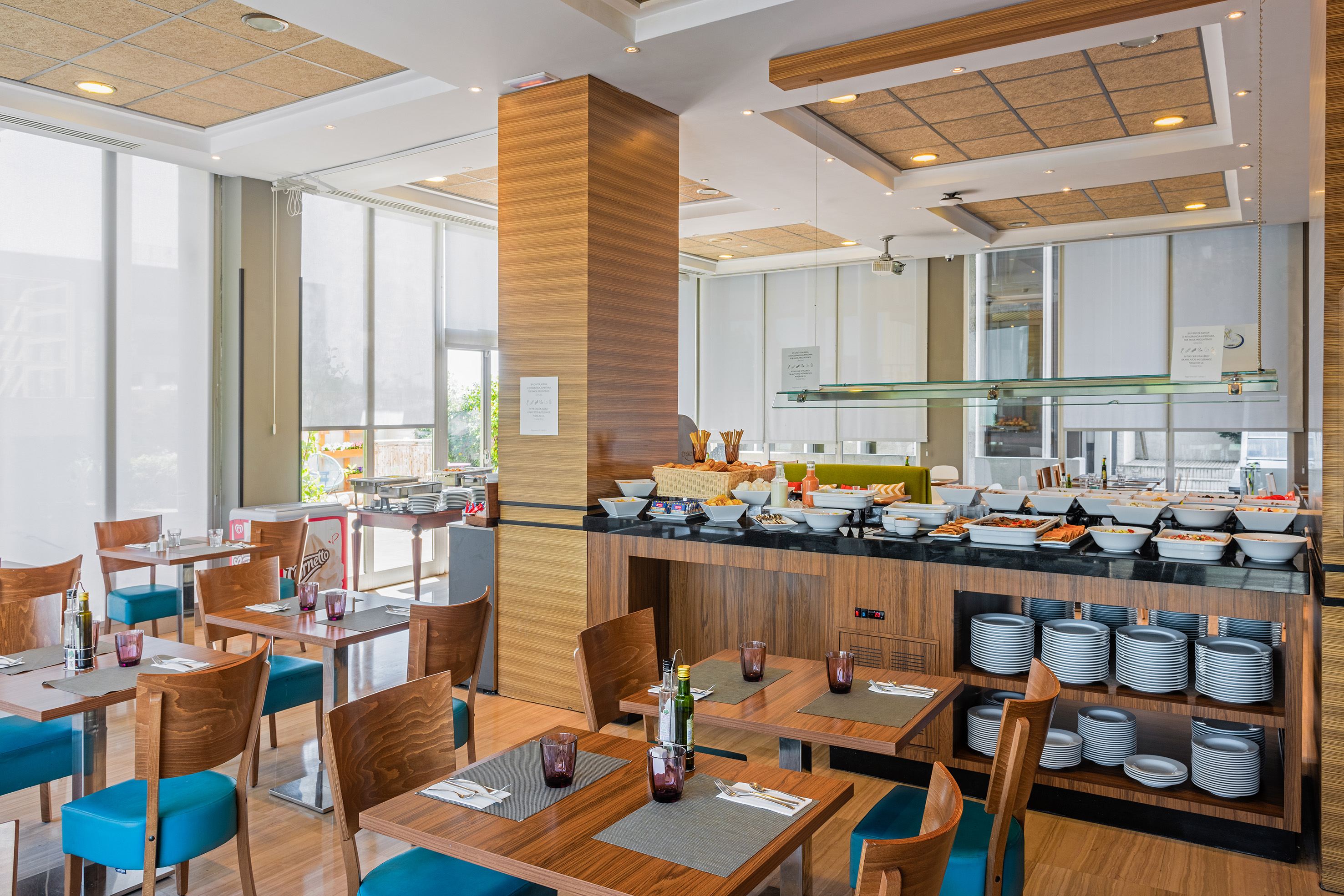 Bright hotel dining room with buffet, wooden tables, and blue chairs.