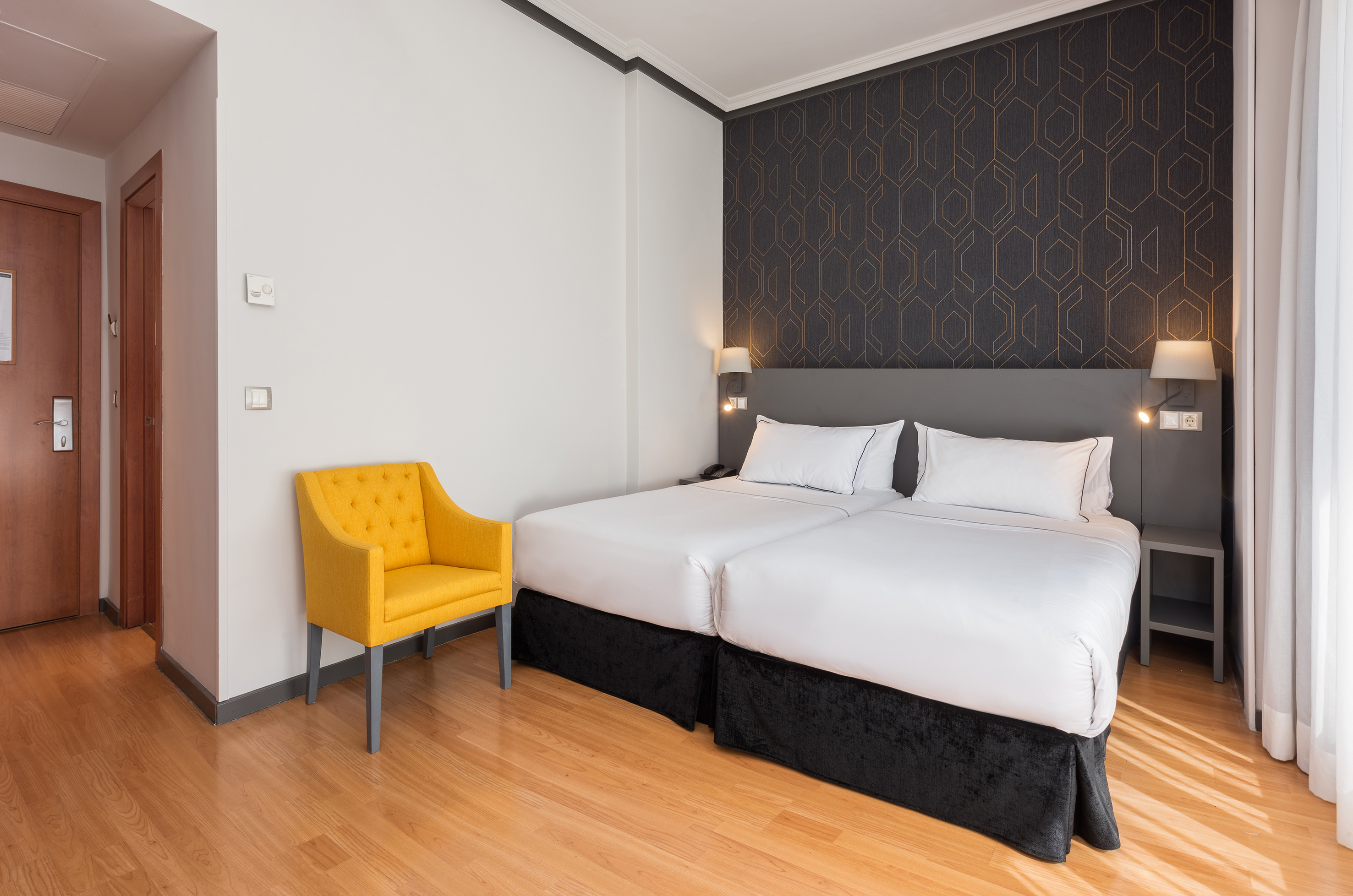 Modern hotel bedroom with twin beds, yellow chair, and geometric accent wall.