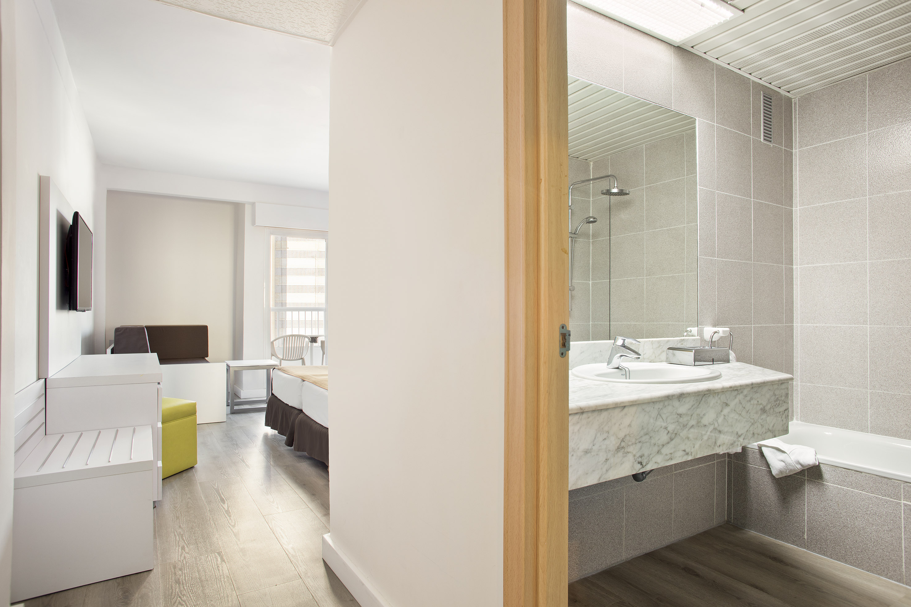 Modern hotel room and bathroom with wood floor, bed, vanity, shower, and tub.