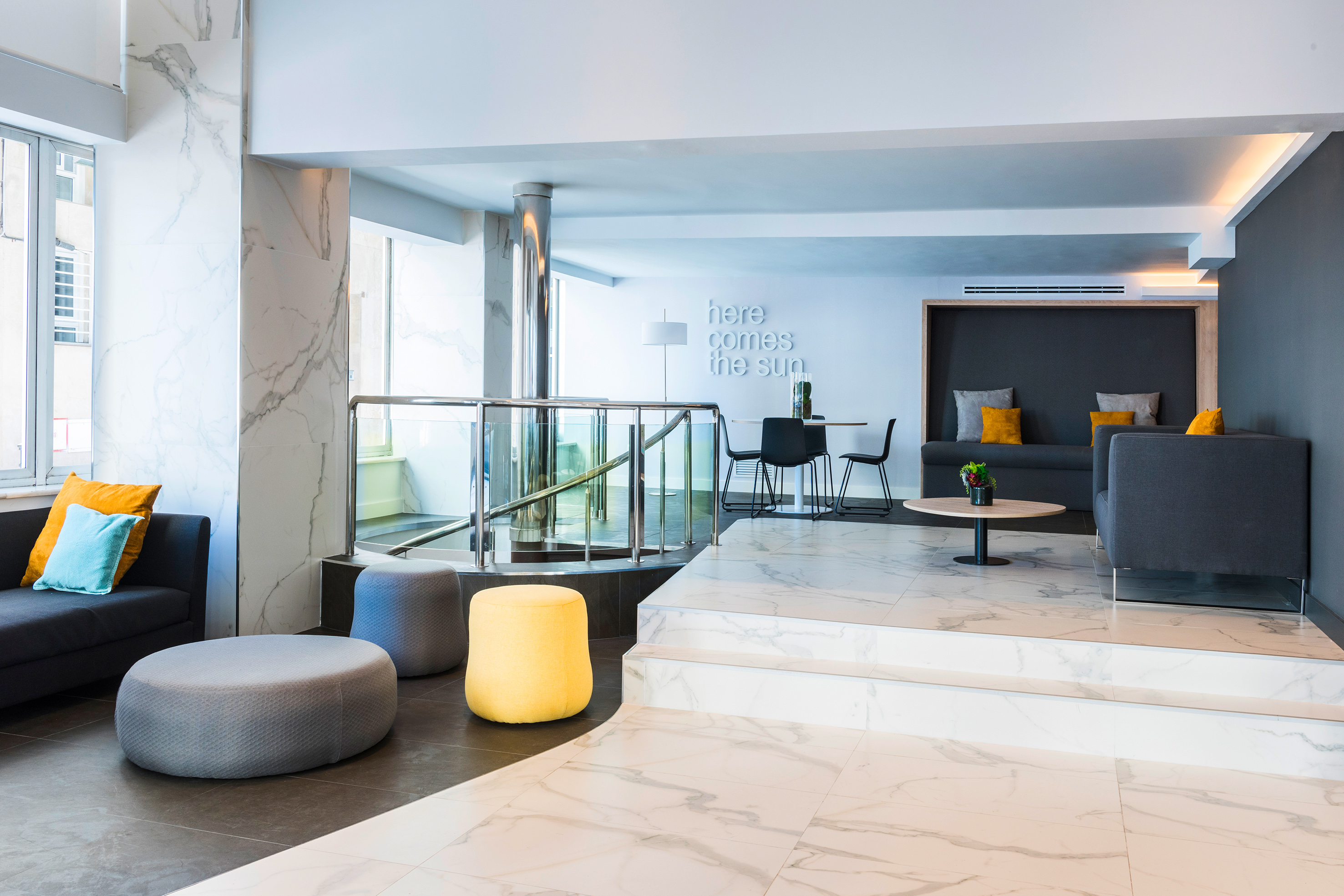 Modern hotel lounge with marble, grey sofas, yellow ottomans, and stylish decor.
