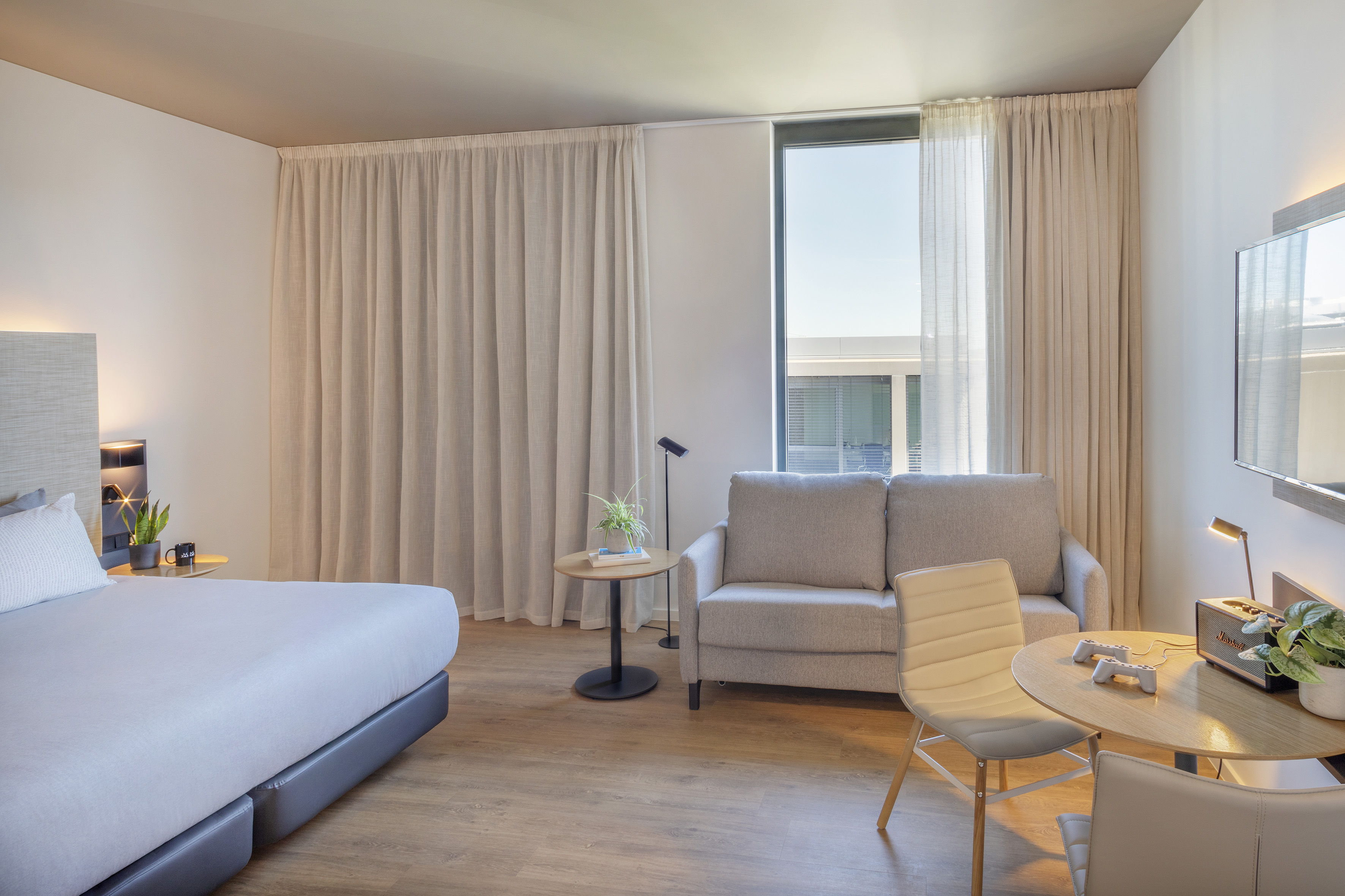 Contemporary hotel room with bed, sofa, dining area, and large window.