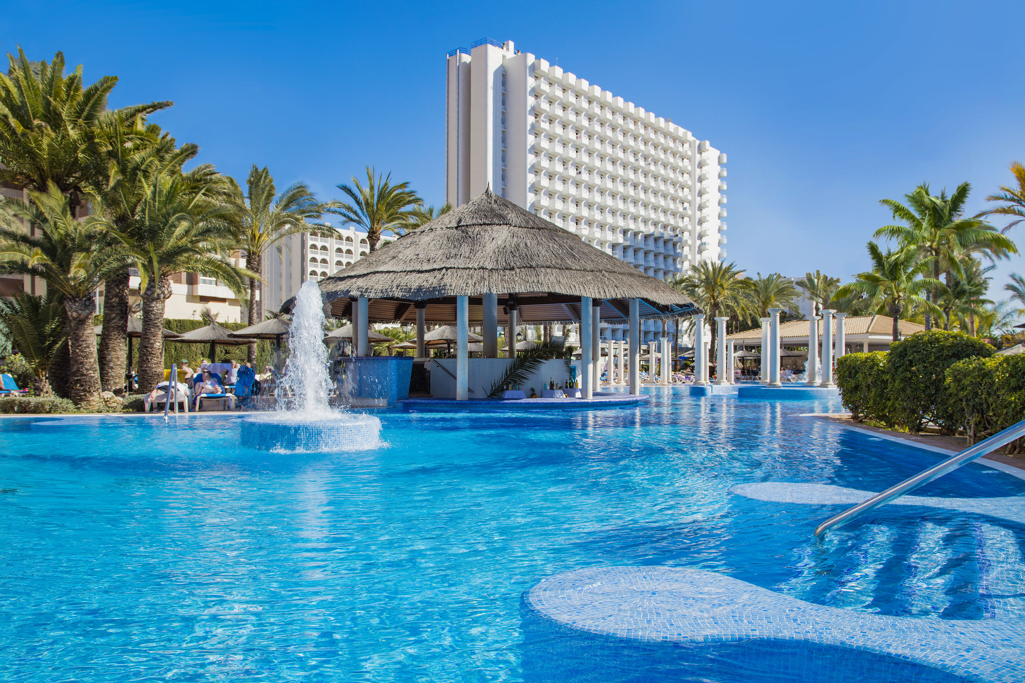 Sunny resort pool, swim-up bar, fountain, palm trees, and hotel building.