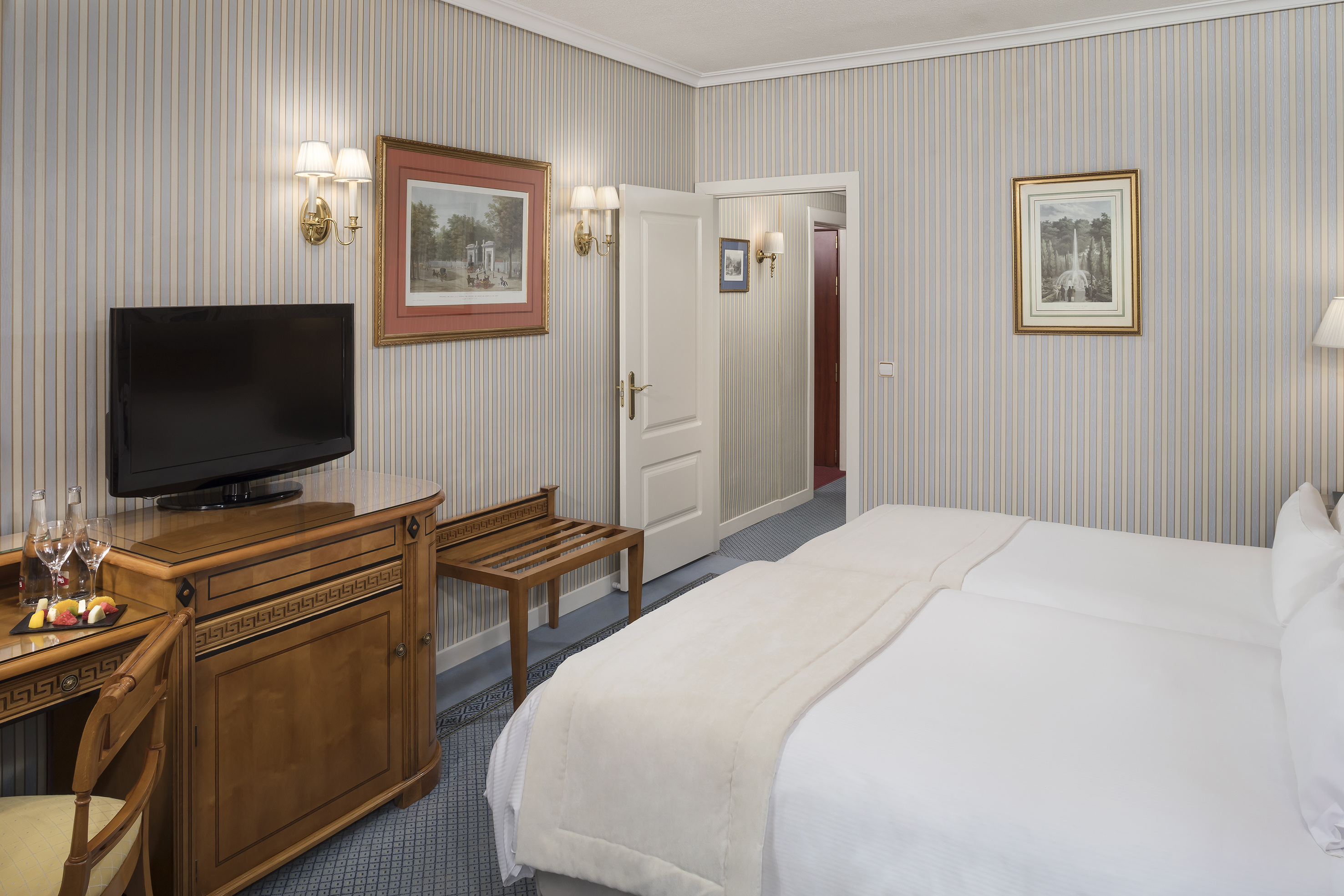 Hotel room with twin beds, striped walls, wooden furniture, TV, and art.