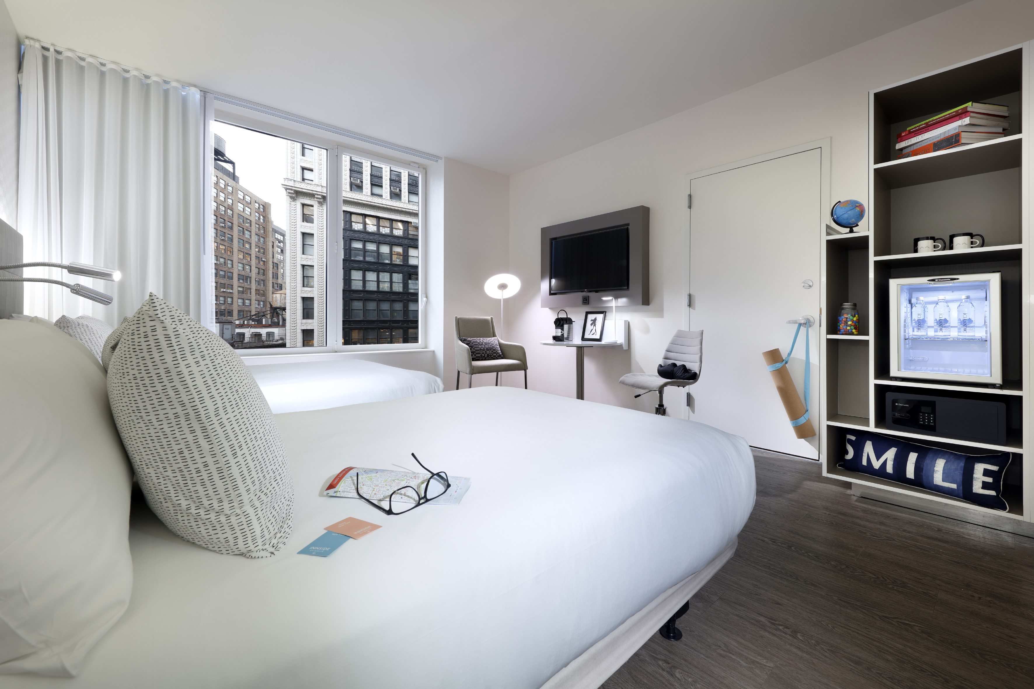 Bright hotel room with two beds, city view, wall-mounted TV, and mini-fridge.