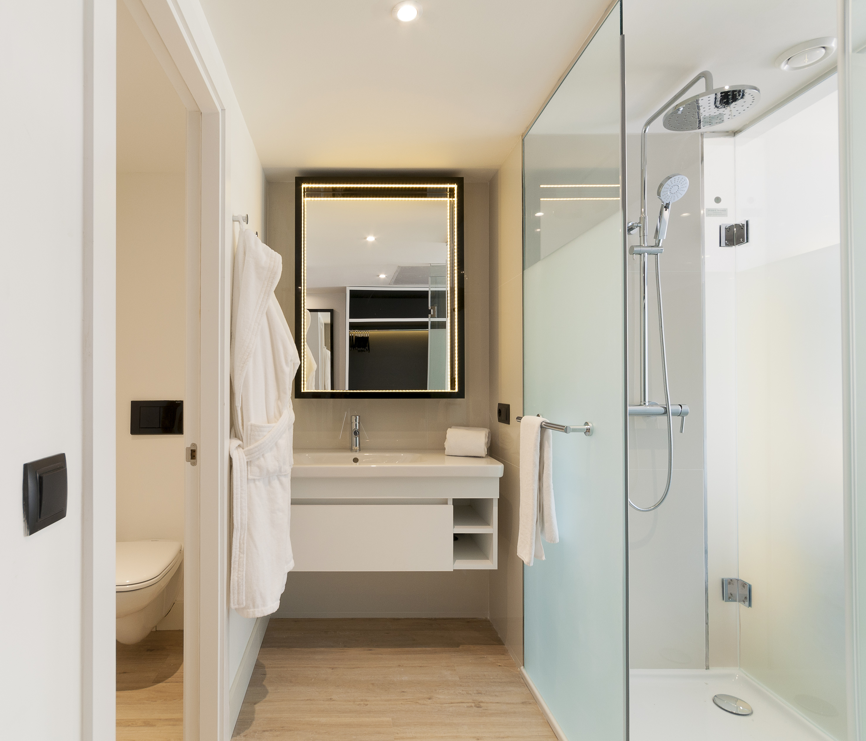 Modern bathroom with glass shower, illuminated mirror, floating vanity, and toilet area.