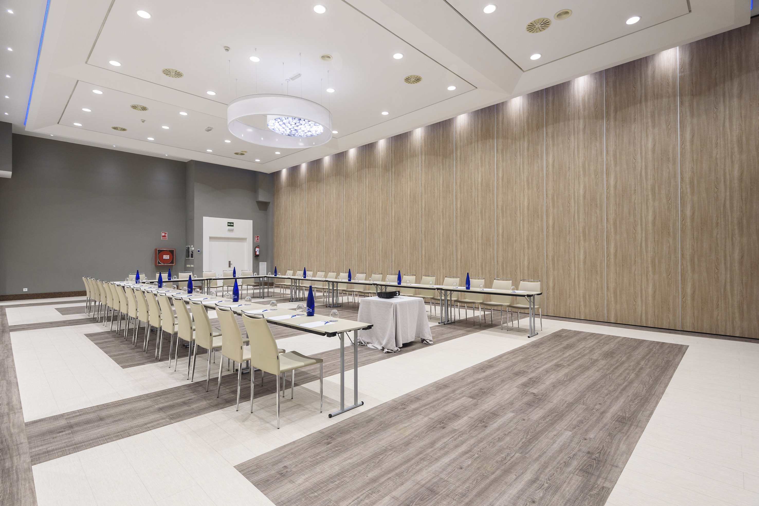 Modern hotel conference room with horseshoe tables, wood walls, and projector setup.