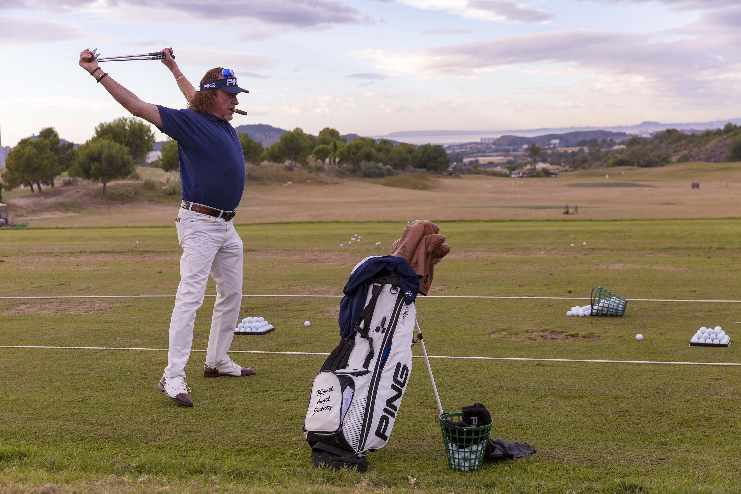 Golfer stretching with golf clubs on a driving range, cigar, Ping bag.
