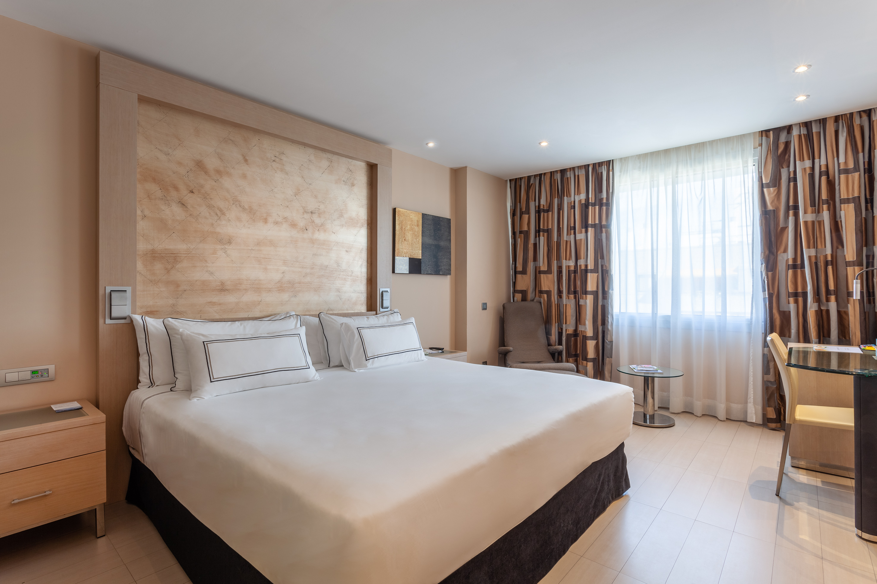 Modern hotel bedroom with king bed, wood headboard, curtains, and desk.