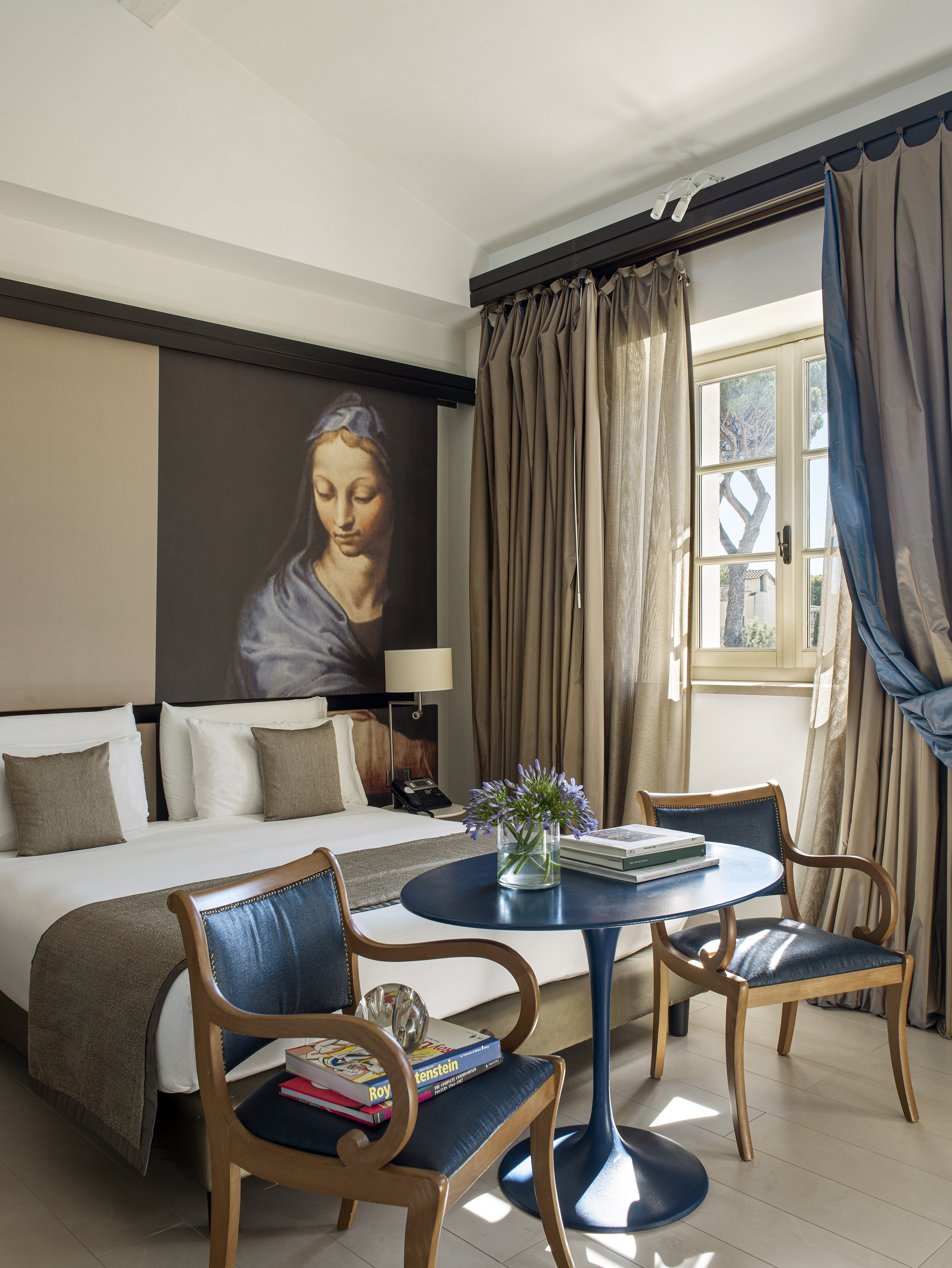 Hotel room with bed, classical art, blue table, chairs, and sunny window.