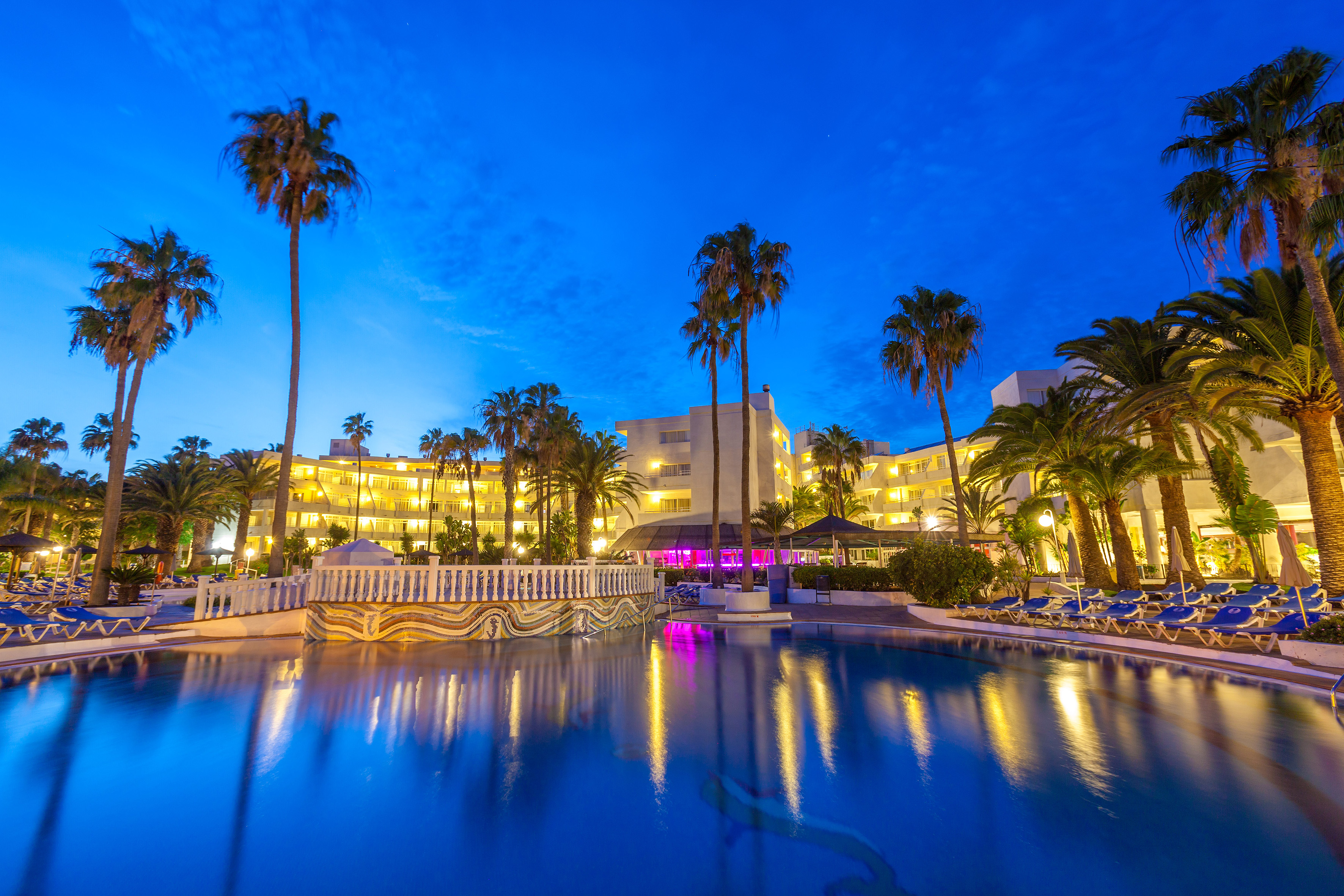 Hotel resort pool at dusk with illuminated buildings and palm trees.