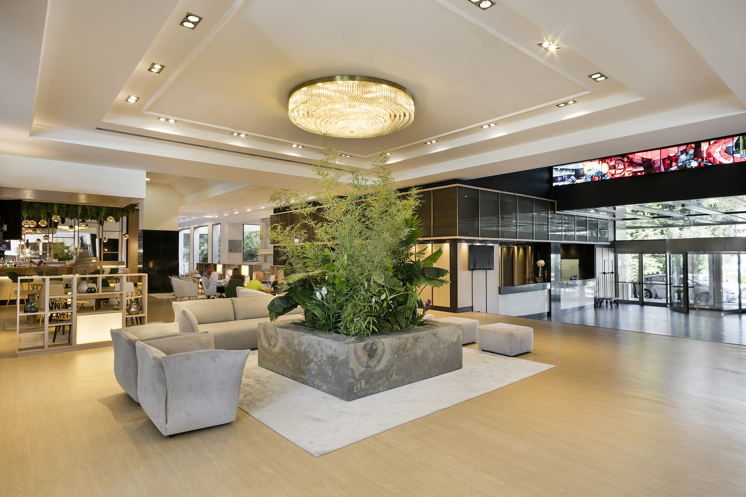 Modern hotel lobby with chandelier, lounge seating, reception, and lush plants.
