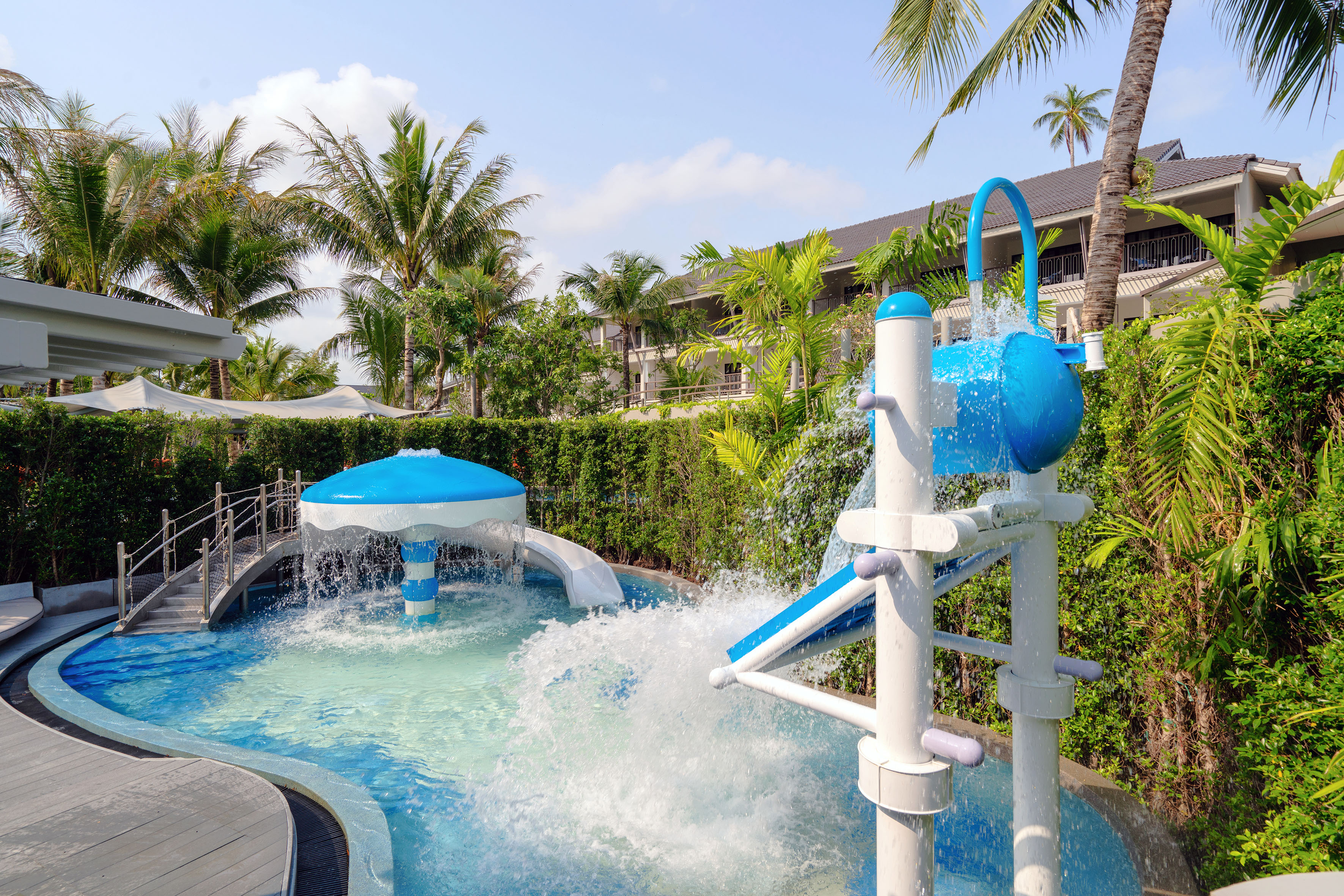 Tropical resort kids' splash pad with blue water features, slides, and palm trees.
