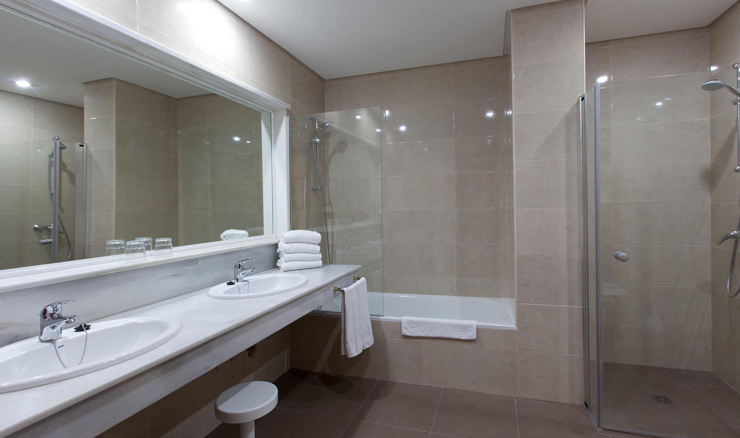 Tiled bathroom with double vanity, large mirror, shower, and bathtub.
