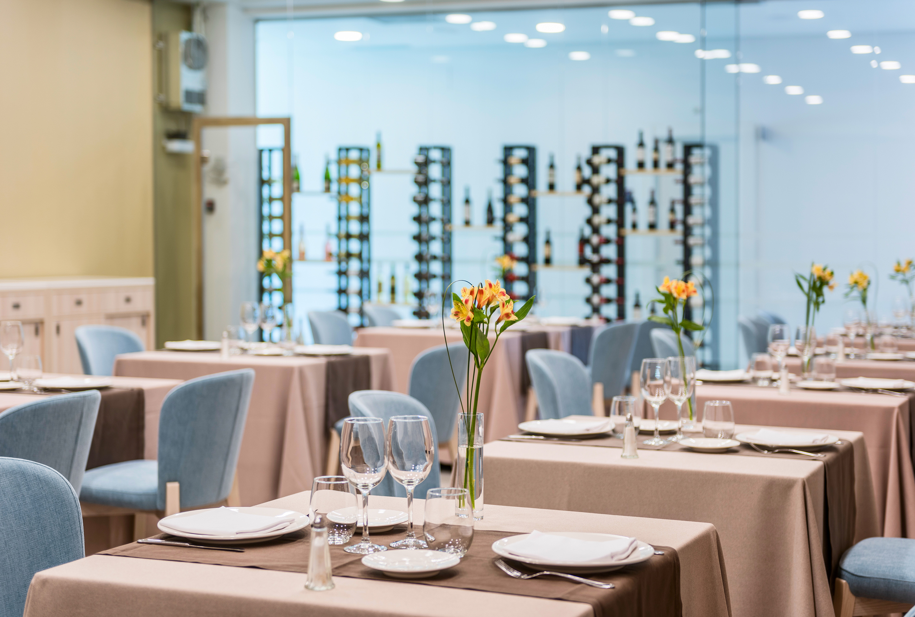 Modern restaurant dining room with set tables, blue chairs, and wine racks.
