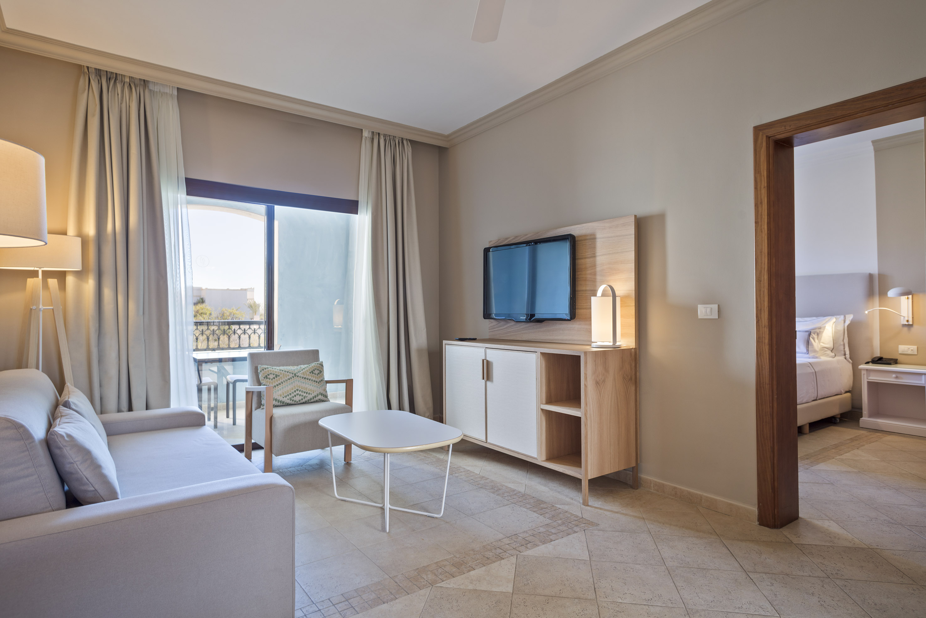 Hotel suite living room with sofa, TV, balcony, and adjacent bedroom.