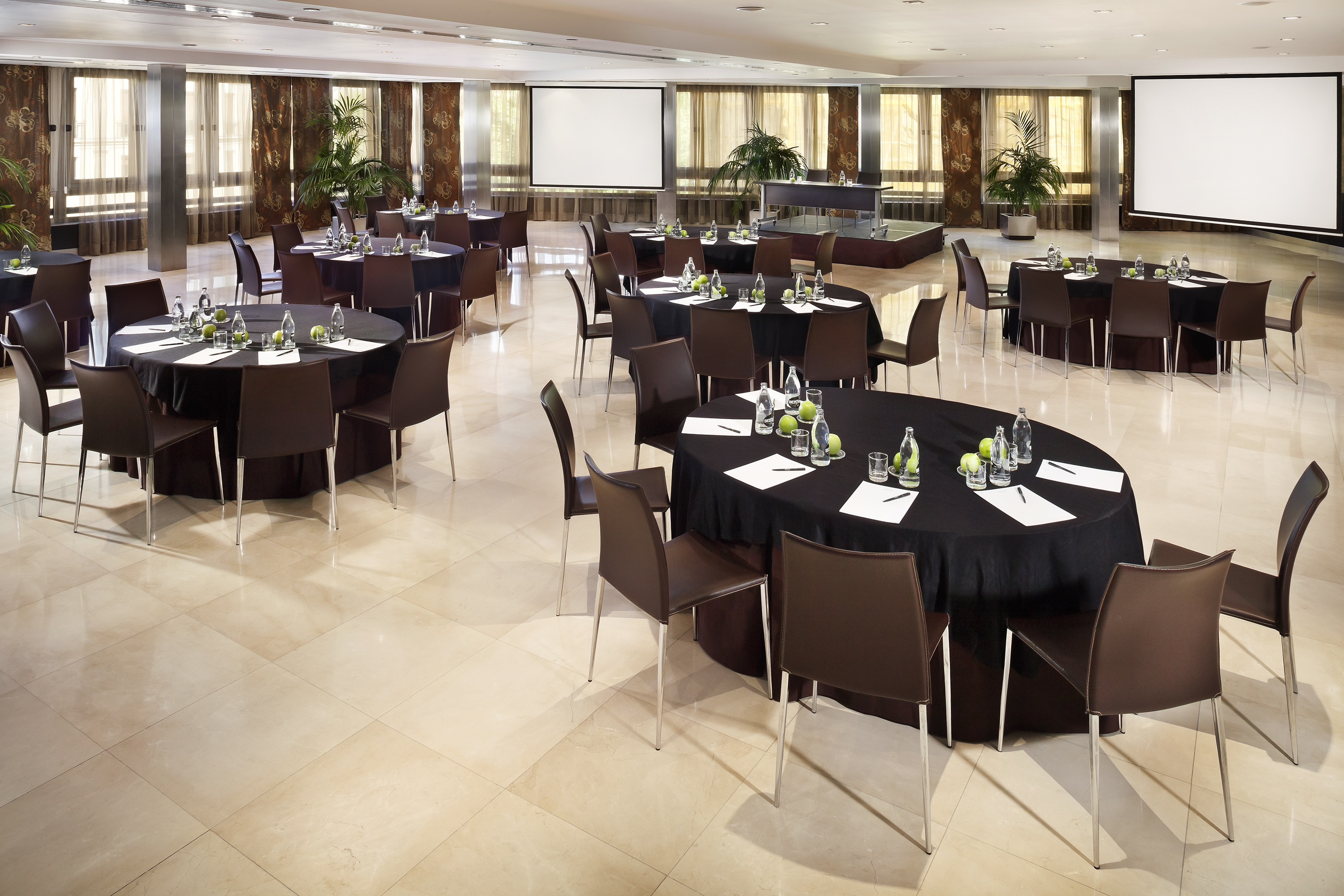 Spacious hotel conference room with multiple round tables, chairs, and two projector screens.