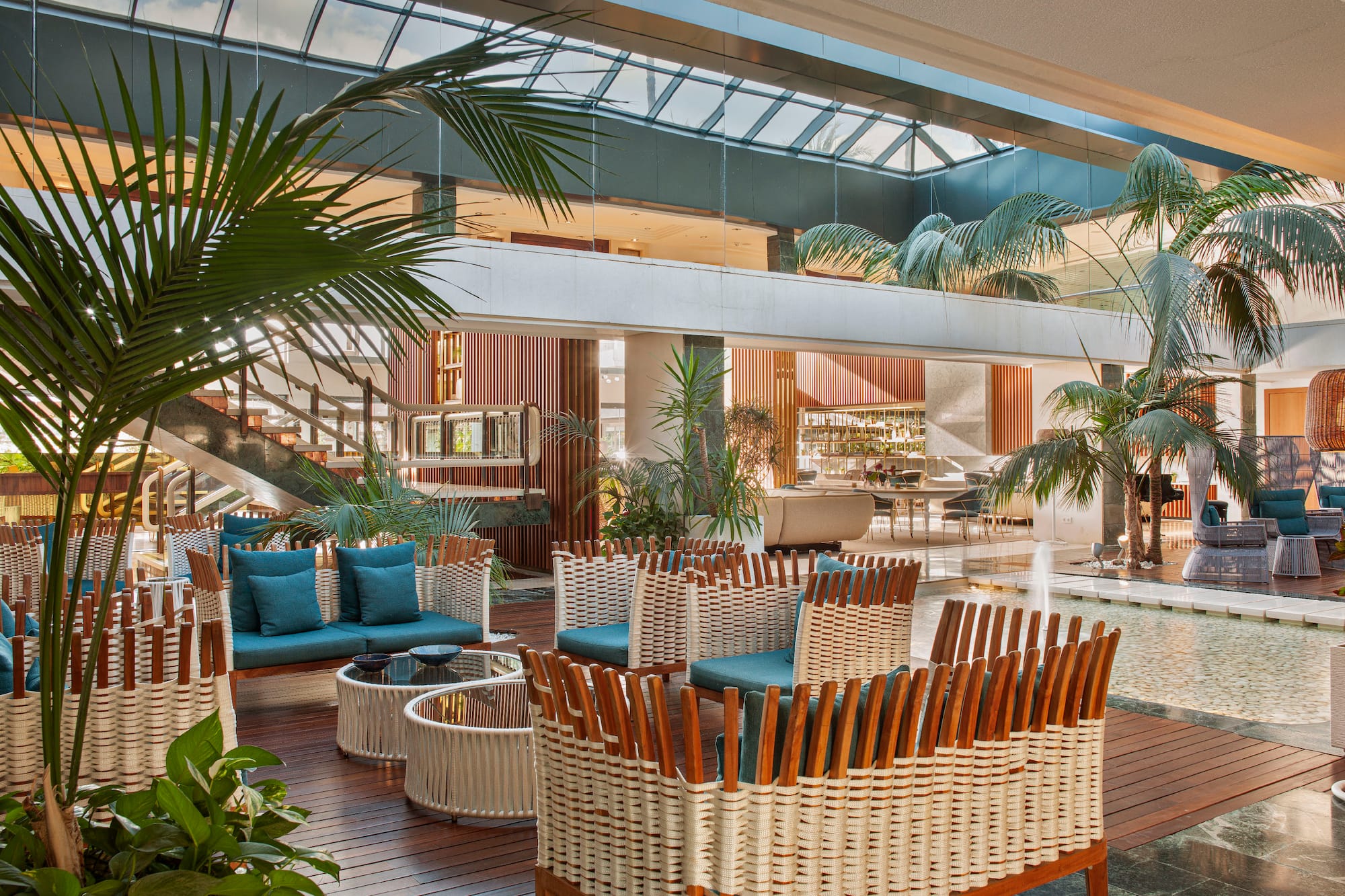 Modern hotel lobby with woven furniture, palm trees, and skylight.