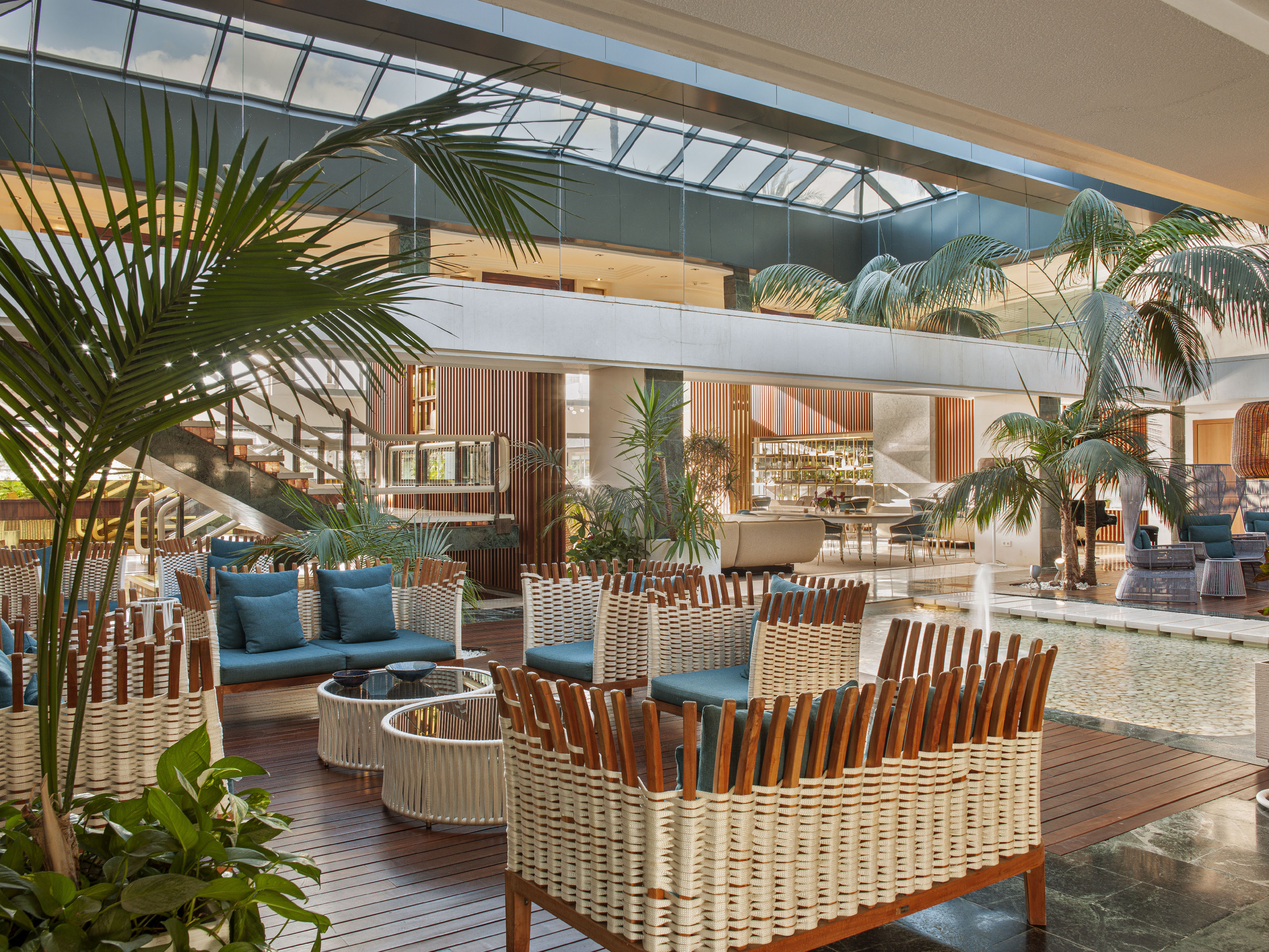 Modern hotel lobby with woven furniture, palm trees, and skylight.
