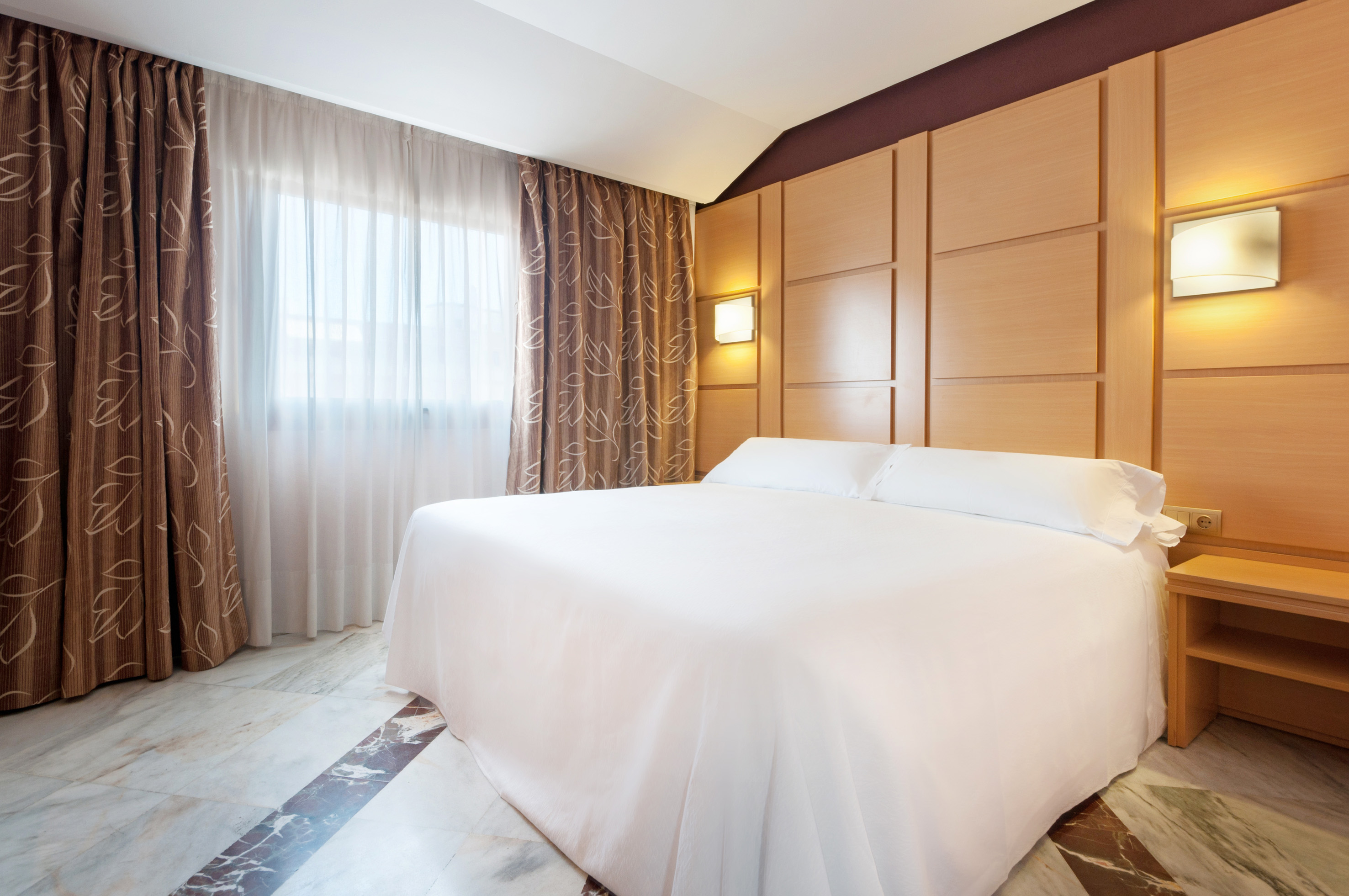 Hotel bedroom with white bed, wood panel headboard, sconce lights, marble floor.