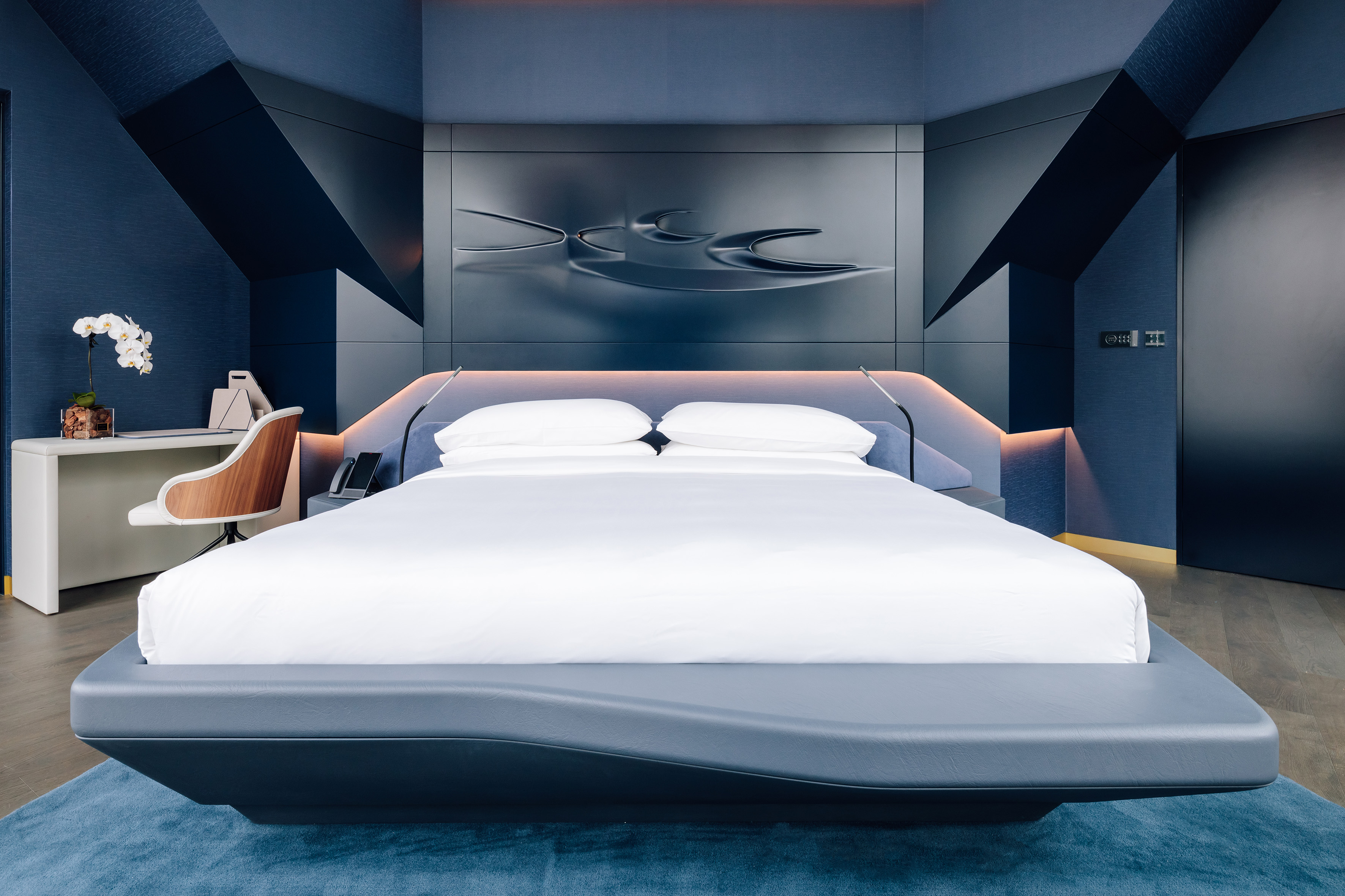 Modern blue luxury hotel bedroom with backlit bed, geometric walls, and sleek desk.