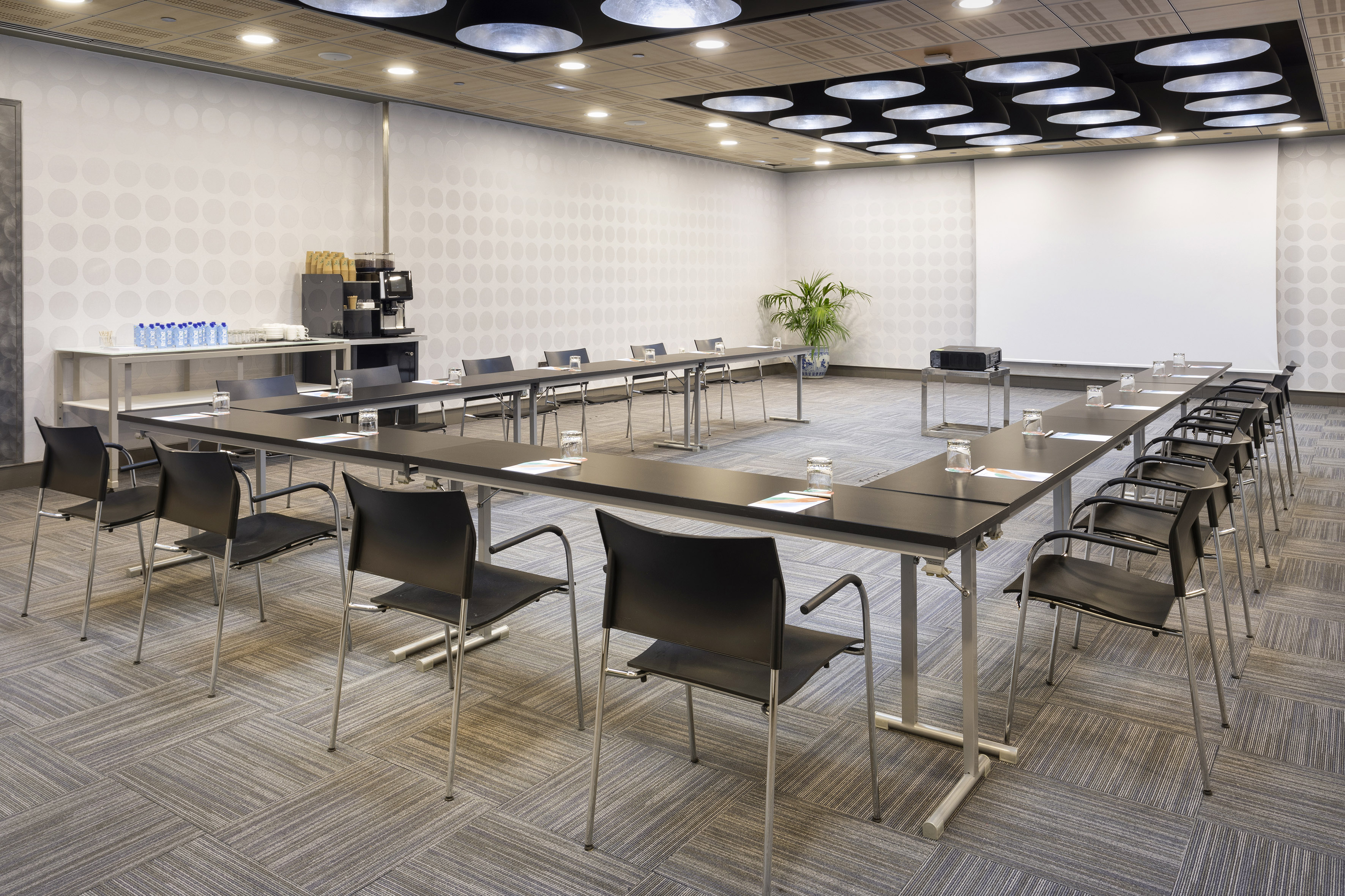 Modern conference room with U-shaped table, projector screen, and coffee station.