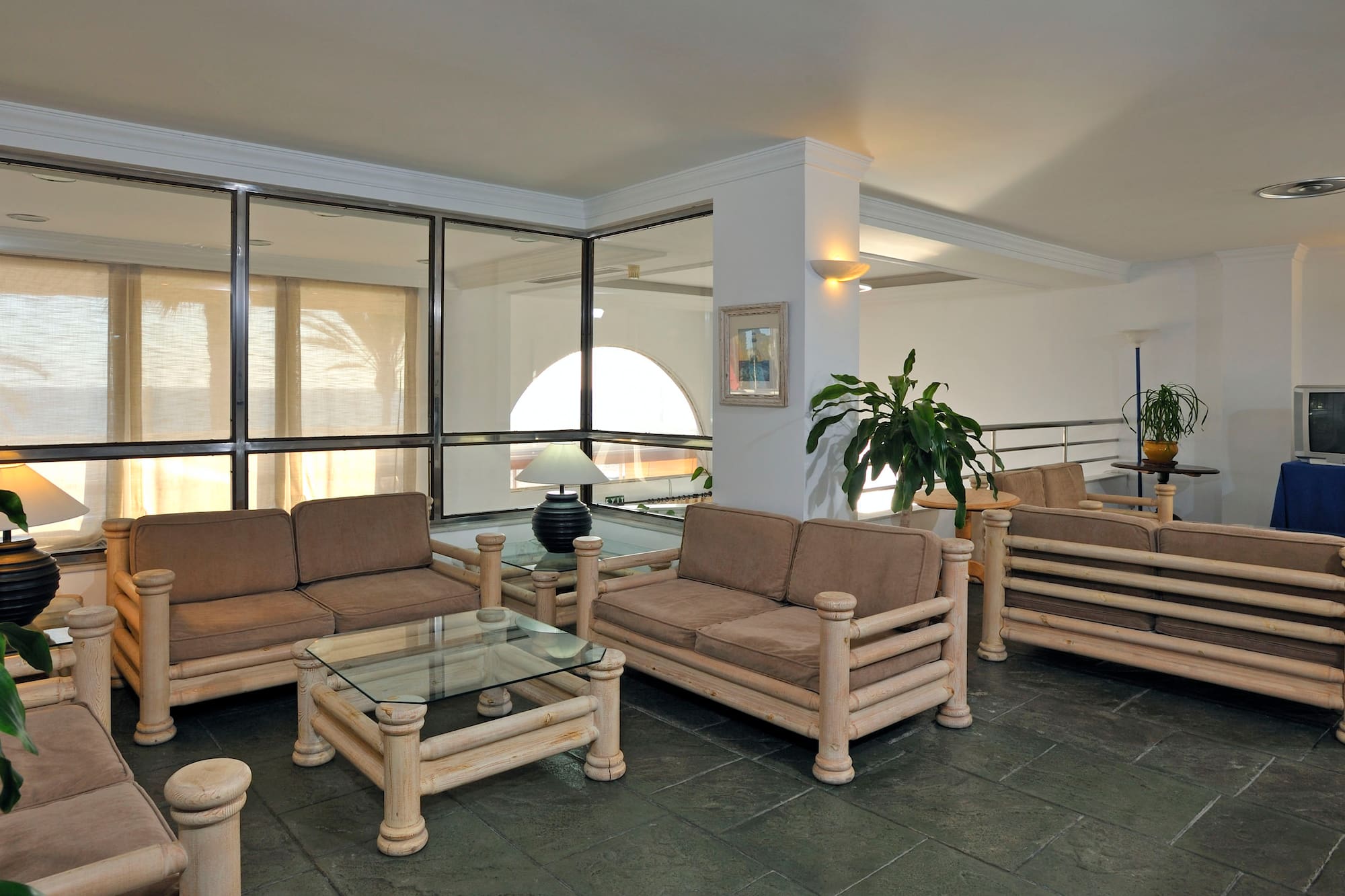 Hotel lobby with light wood sofas, glass tables, potted plants, and large windows.