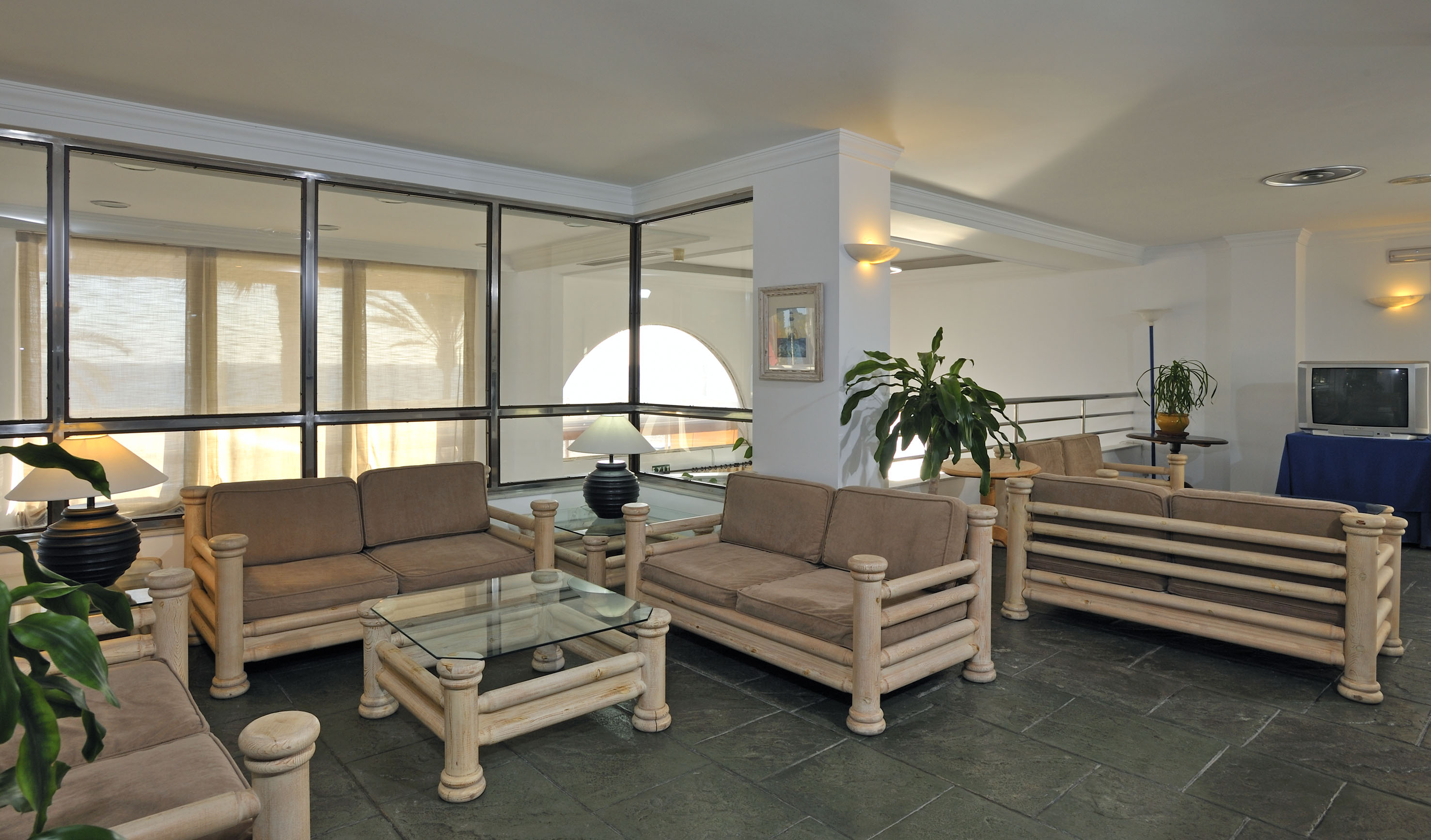 Hotel lobby with light wood sofas, glass tables, potted plants, and large windows.