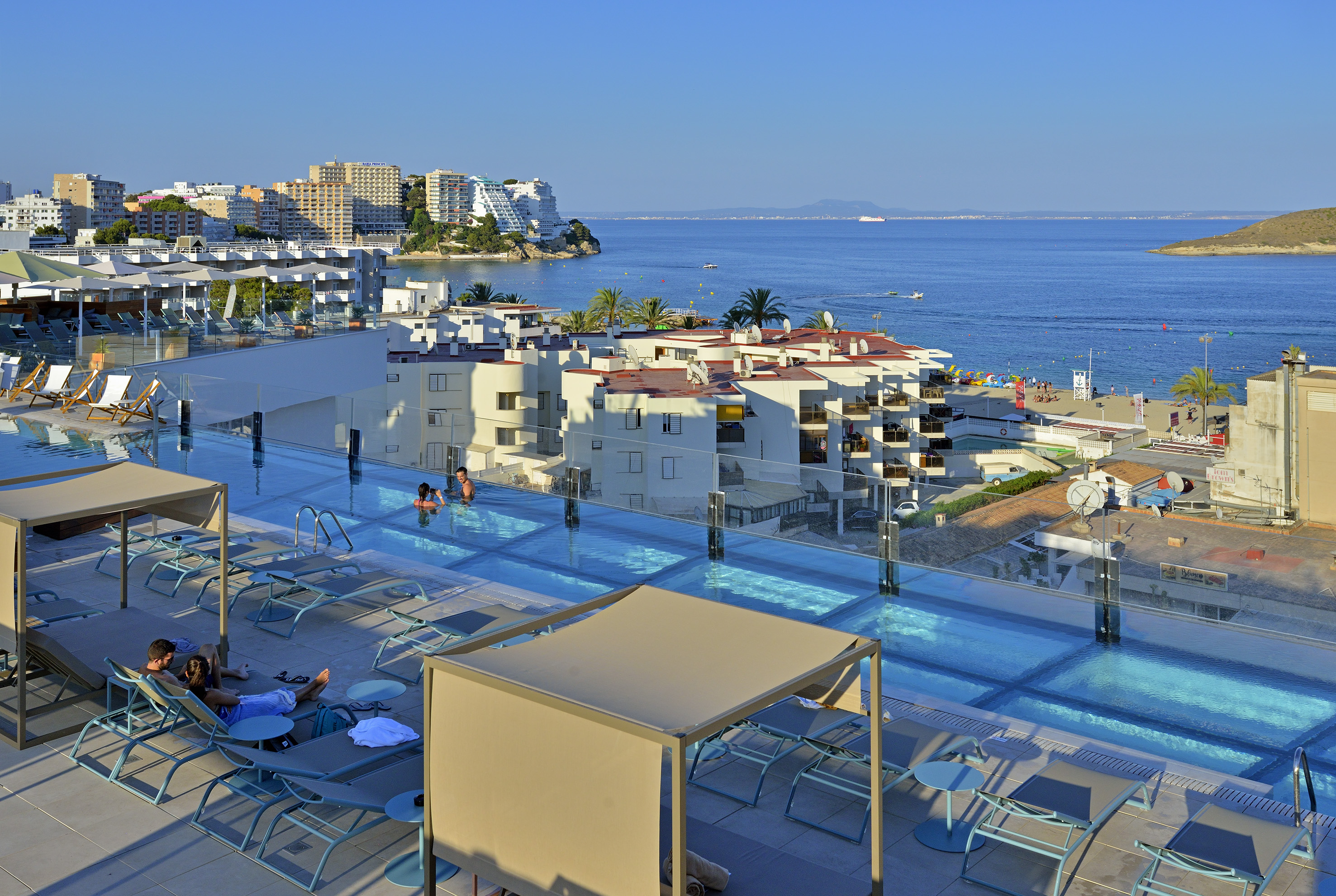 Rooftop infinity pool with ocean view, coastal buildings, and sunbathers.