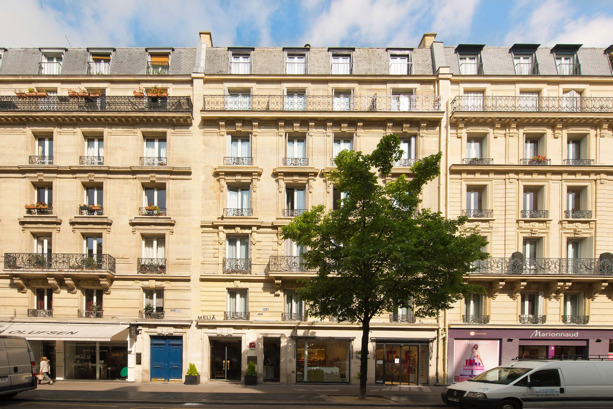 Haussmannian building facade in Paris with storefronts, balconies, and a large tree.