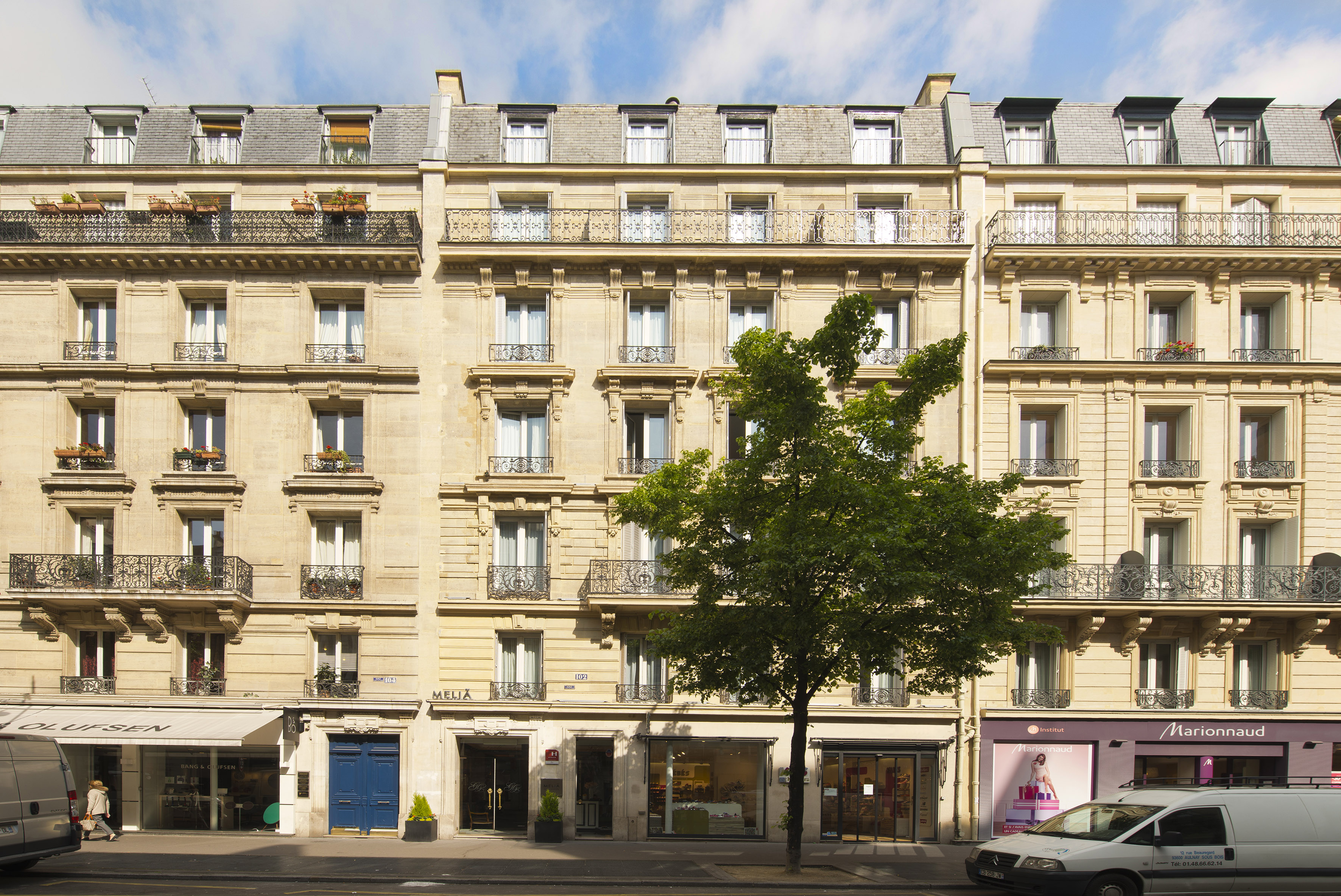 Haussmannian building facade in Paris with storefronts, balconies, and a large tree.