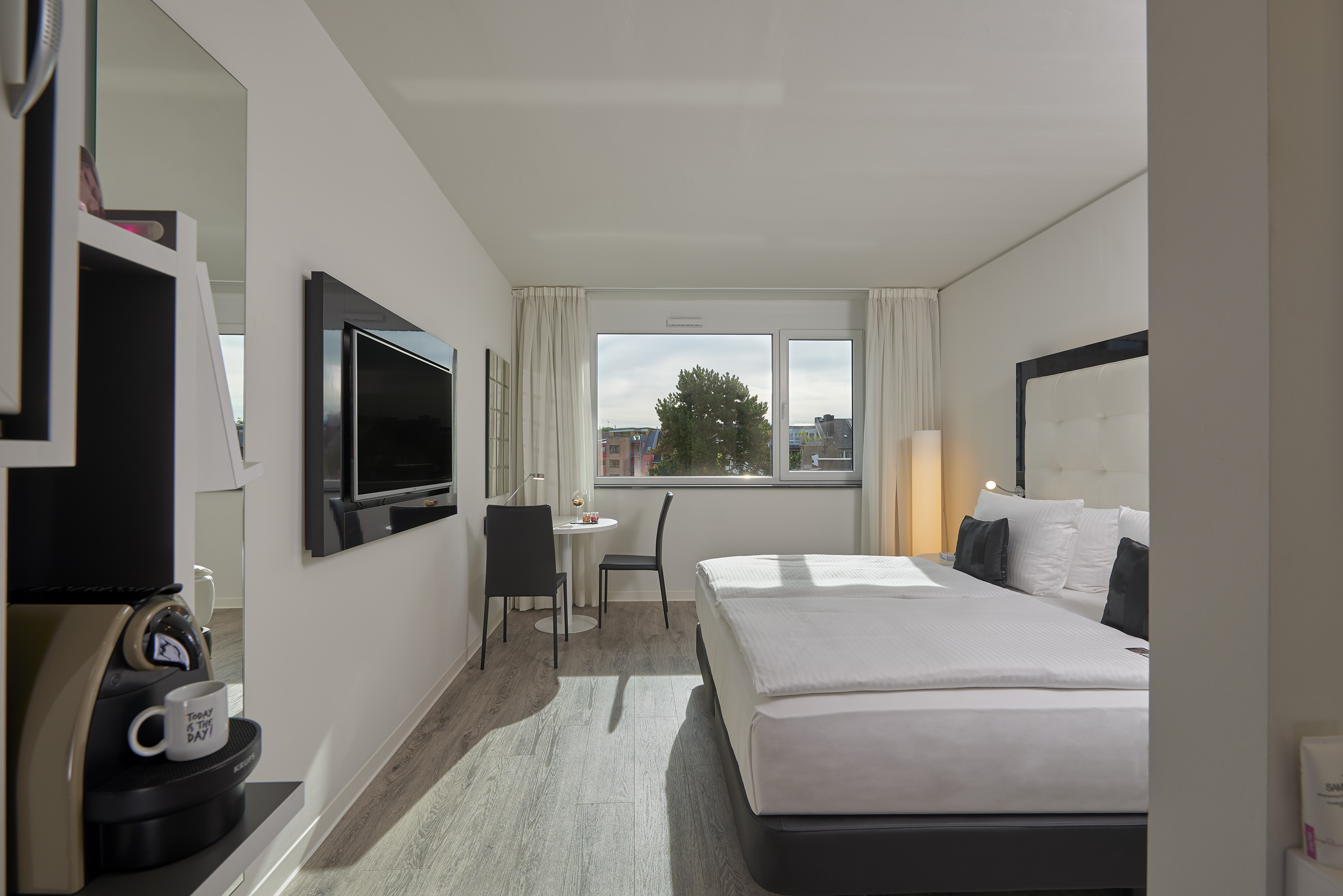 Modern hotel room with queen bed, wall-mounted TV, coffee machine, and city view.