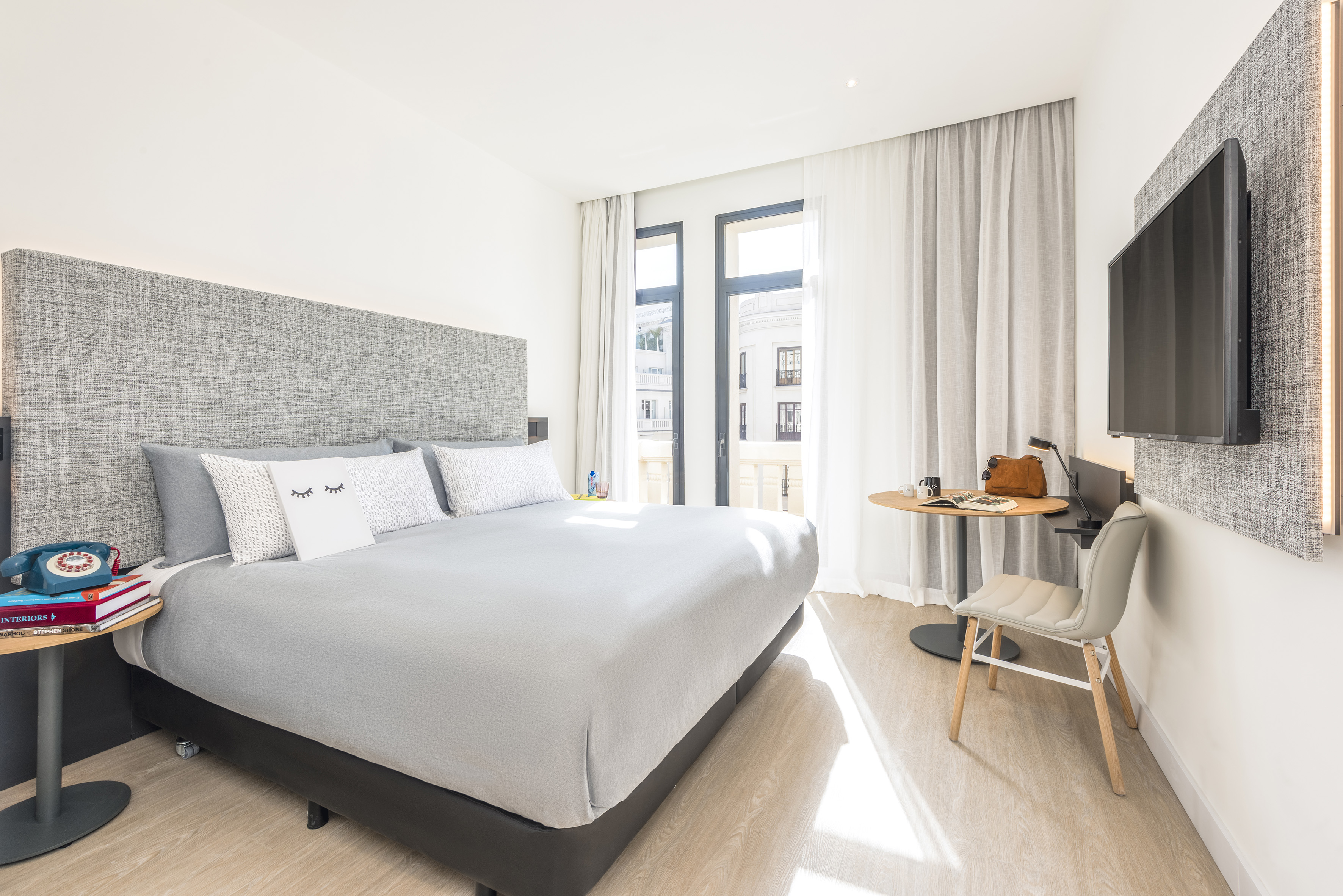 Modern hotel bedroom with large grey bed, balcony access, and flat-screen TV.