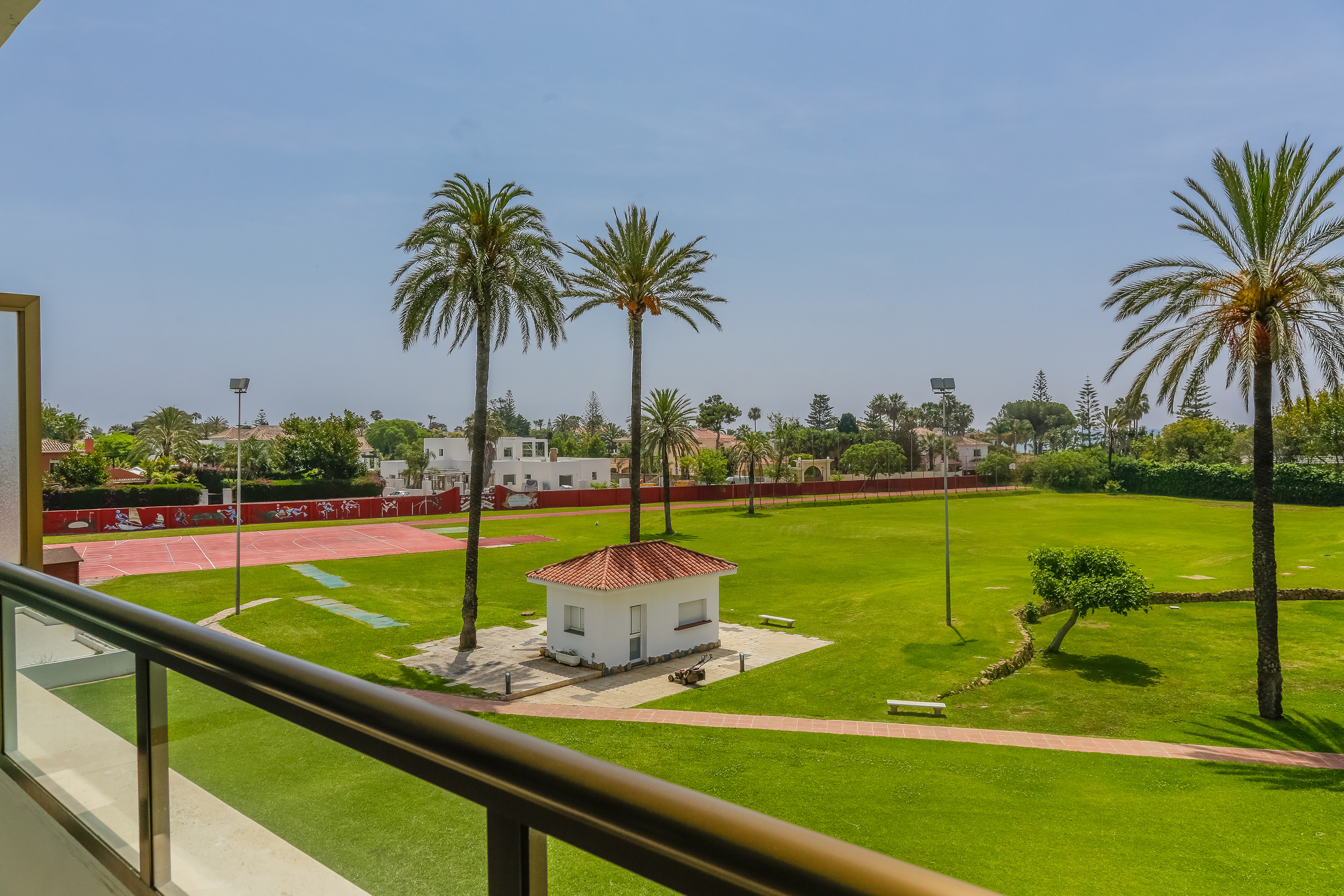Balcony view: resort lawn with palm trees, clubhouse, and red sports court.
