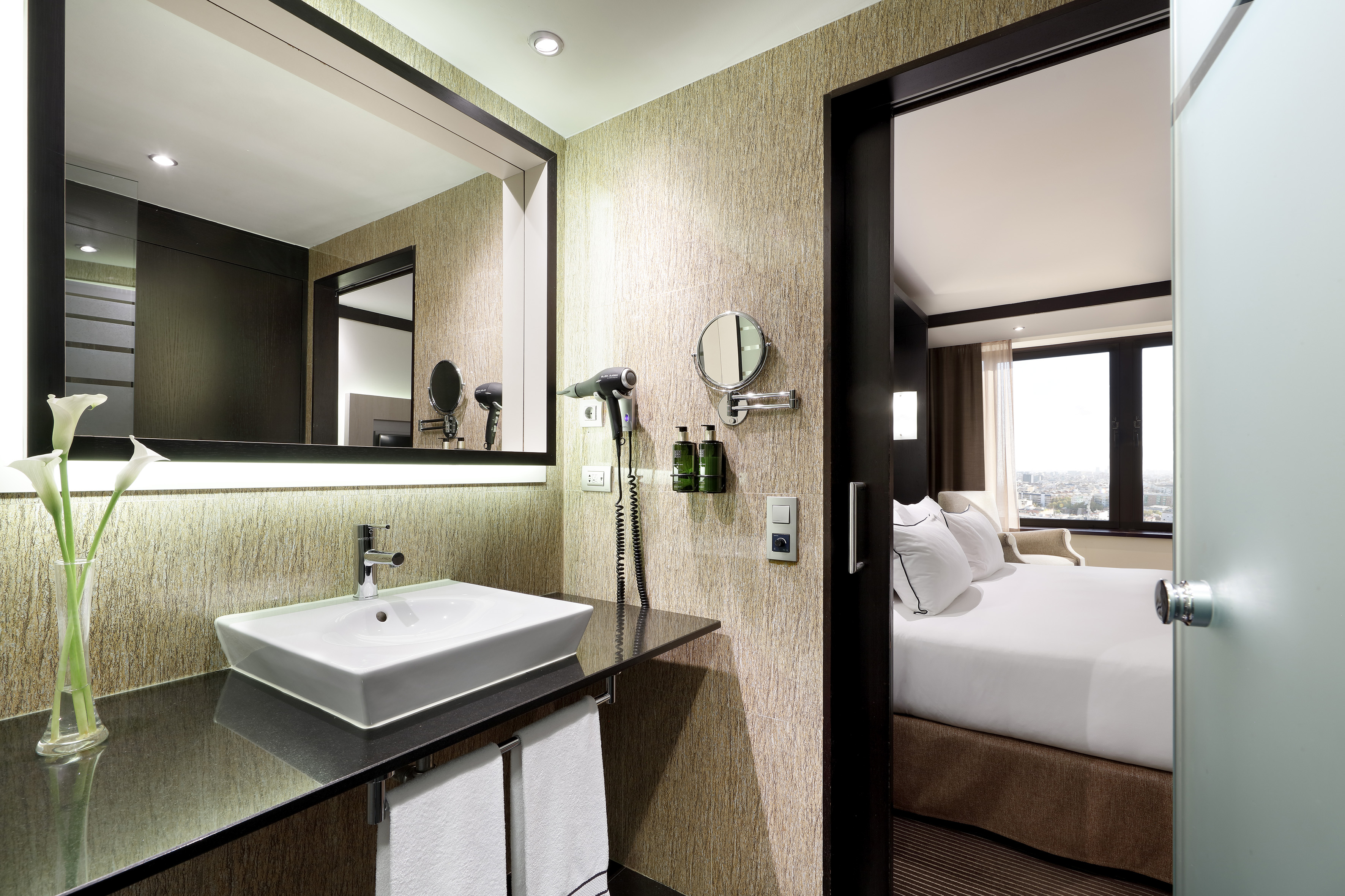 Hotel bathroom vanity with sink, mirror. View into hotel bedroom with city view.