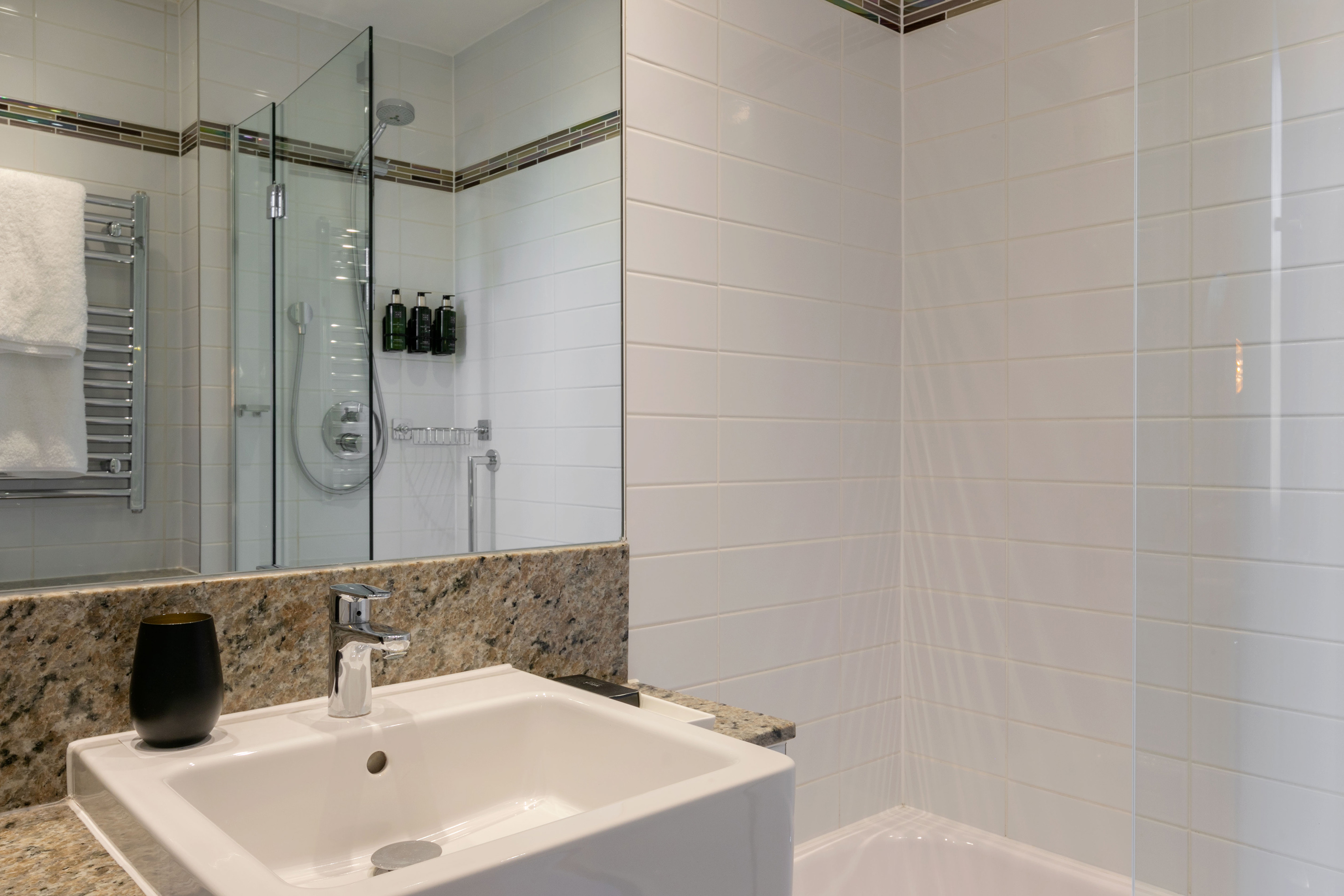 Modern hotel bathroom with white tiles, glass shower, sink, and granite counter.