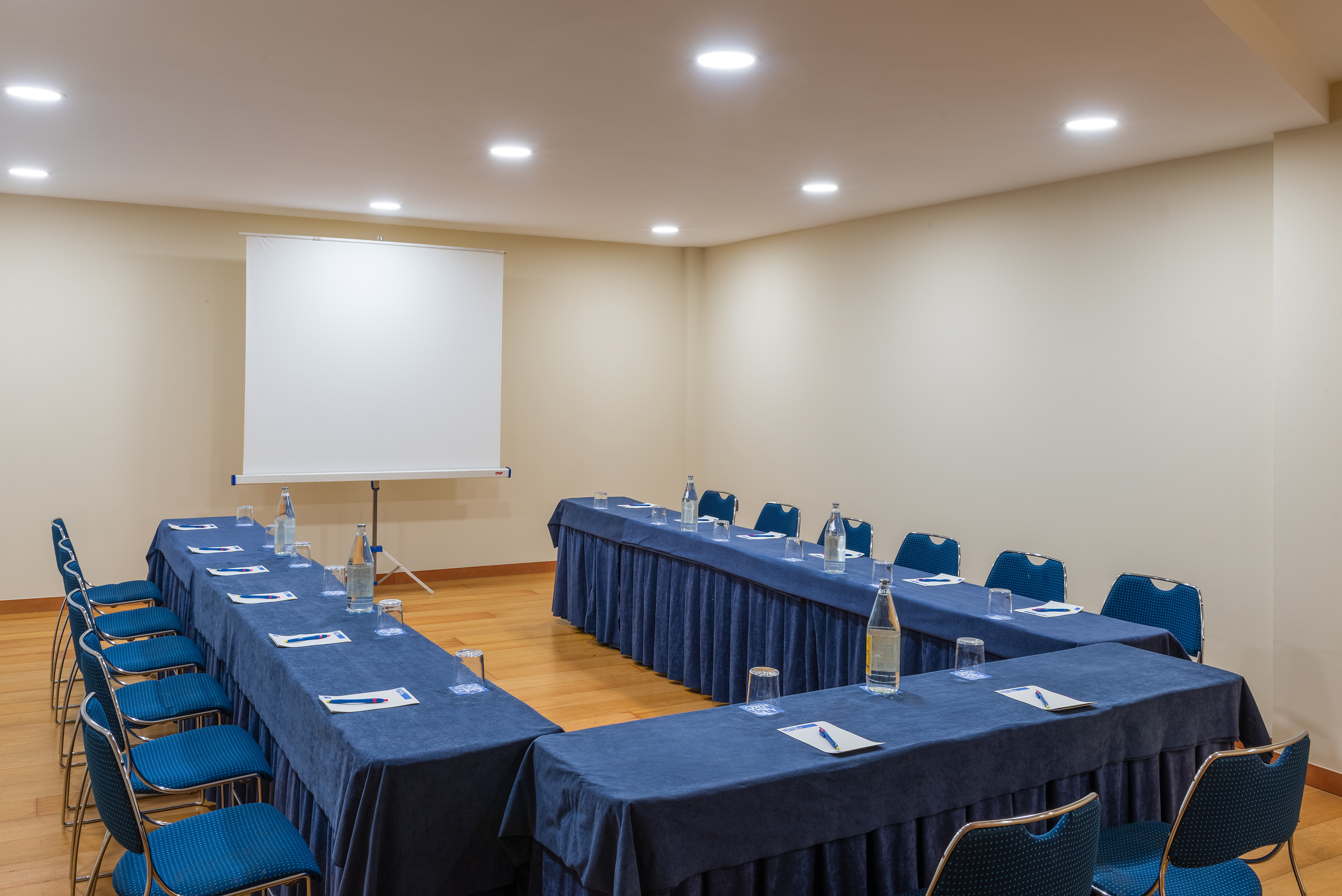 U-shape conference room with blue tables, chairs, water, and projector screen.