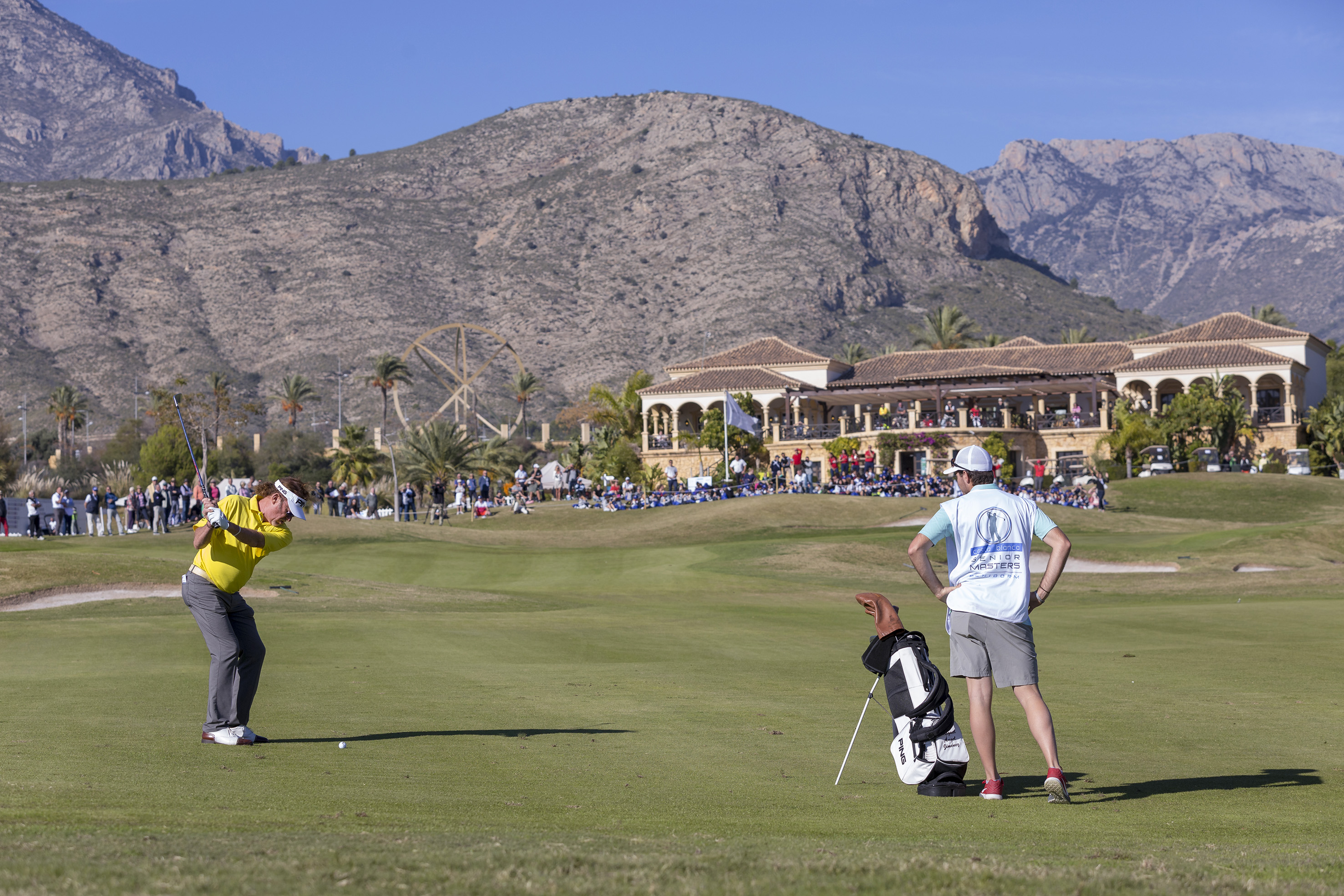 Golfer swings at Senior Masters golf event, caddie, clubhouse, and mountains.