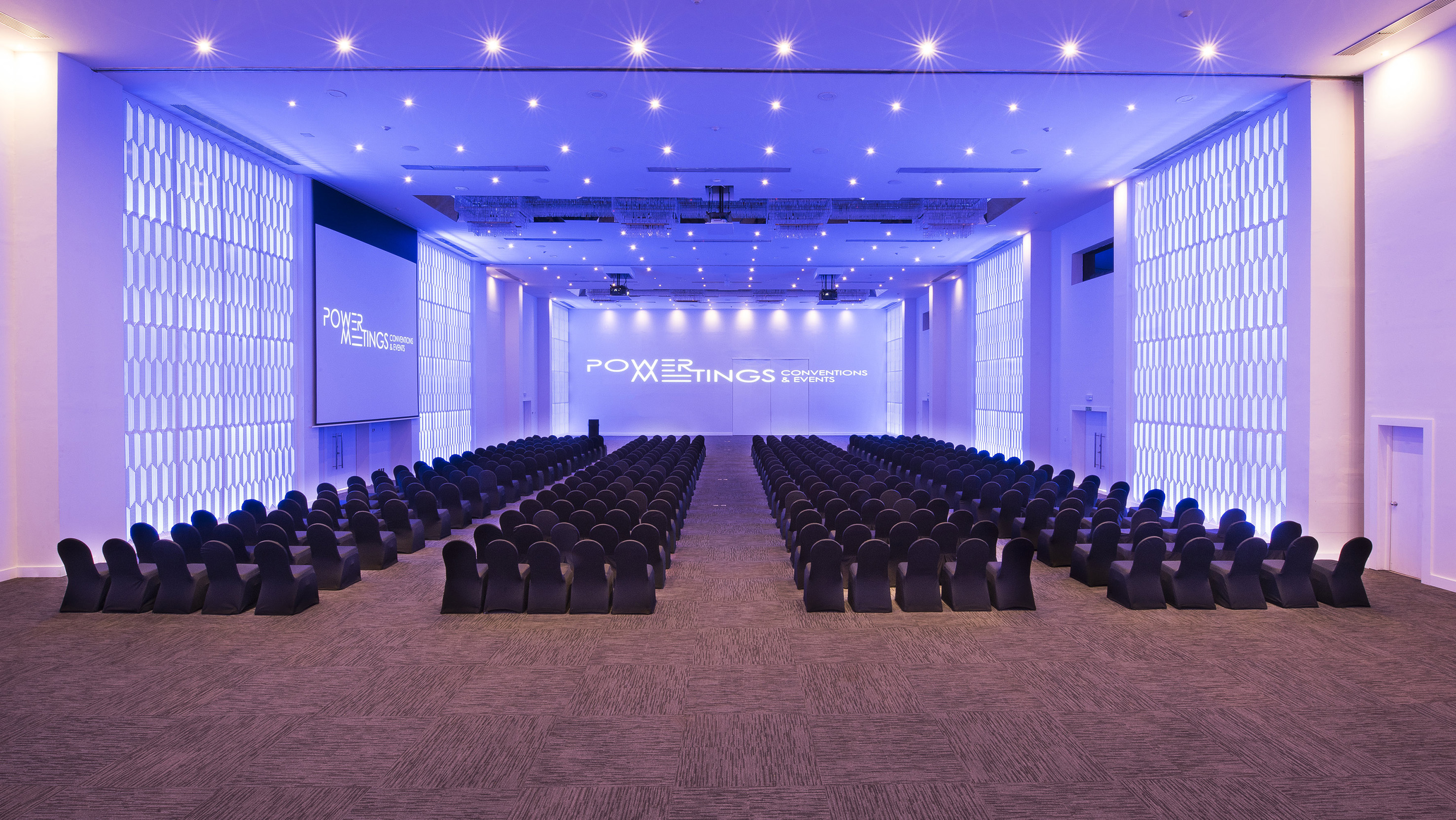 Conference hall with blue lighting, rows of chairs, 'Power Meetings' display.