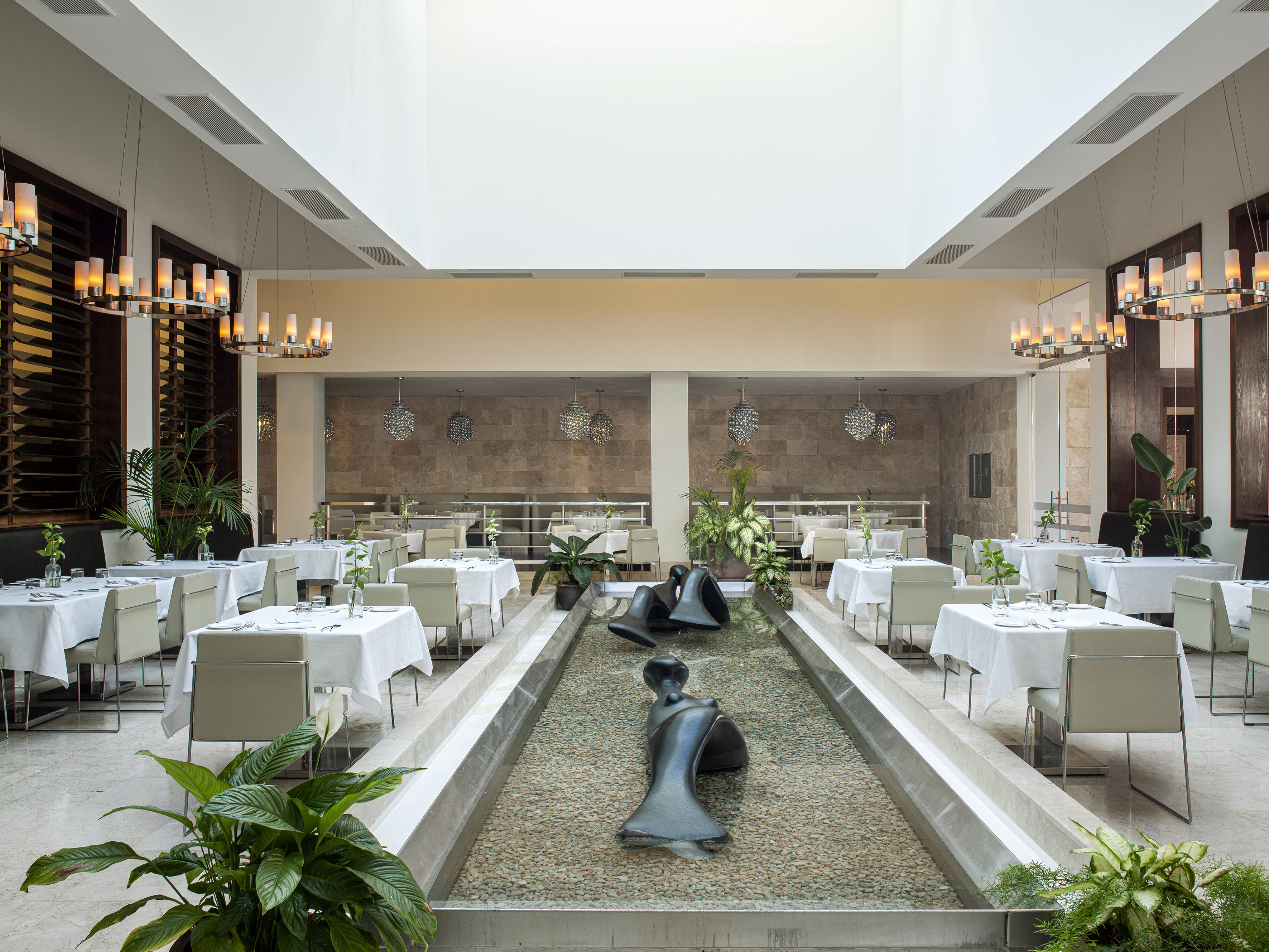 Modern hotel restaurant with white tables, central fountain, and sleek chandeliers.